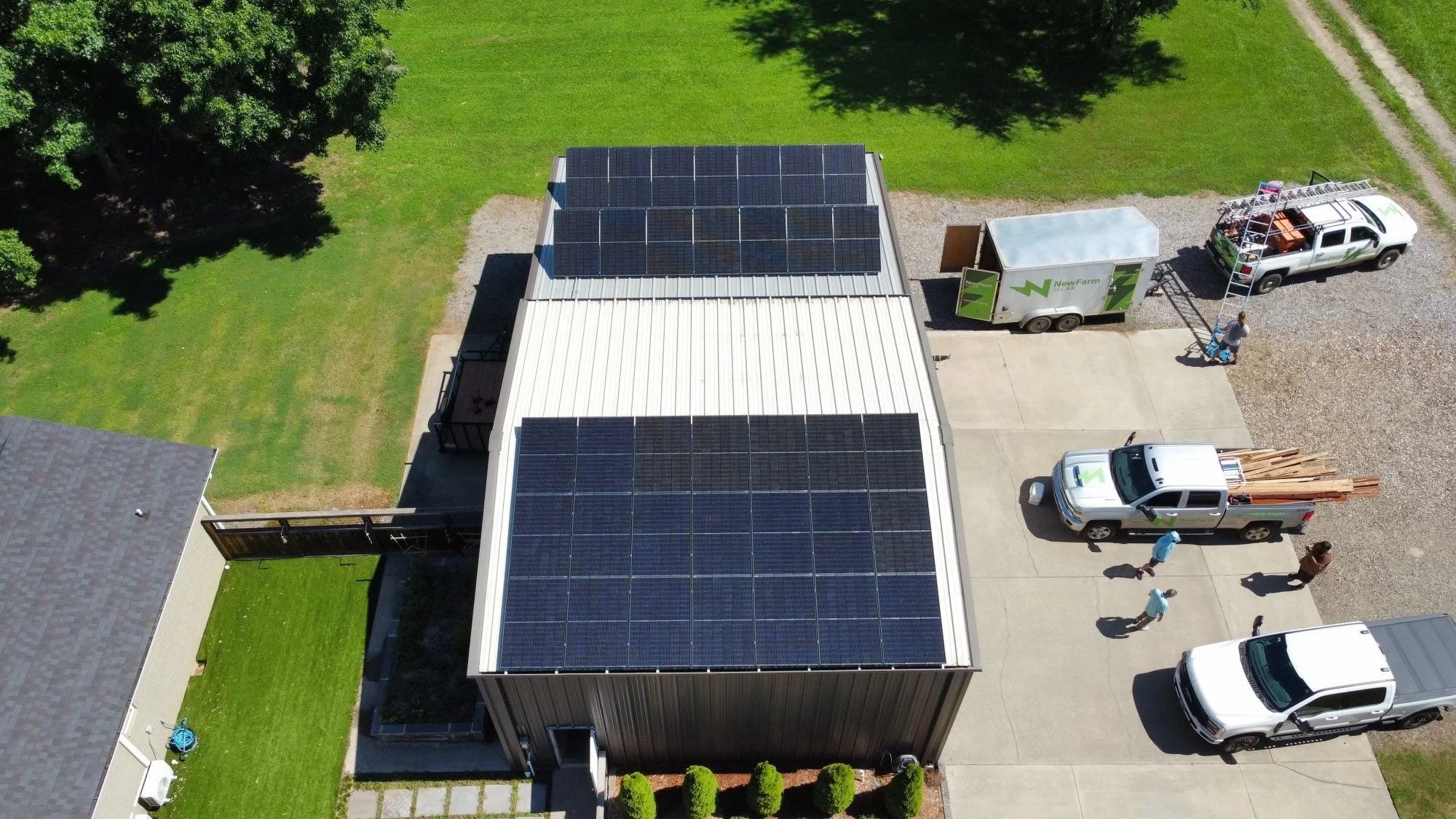 An aerial view of a house with solar panels on the roof, a driveway with cars, a green lawn, and a truck with equipment parked next to the house, with people walking nearby.