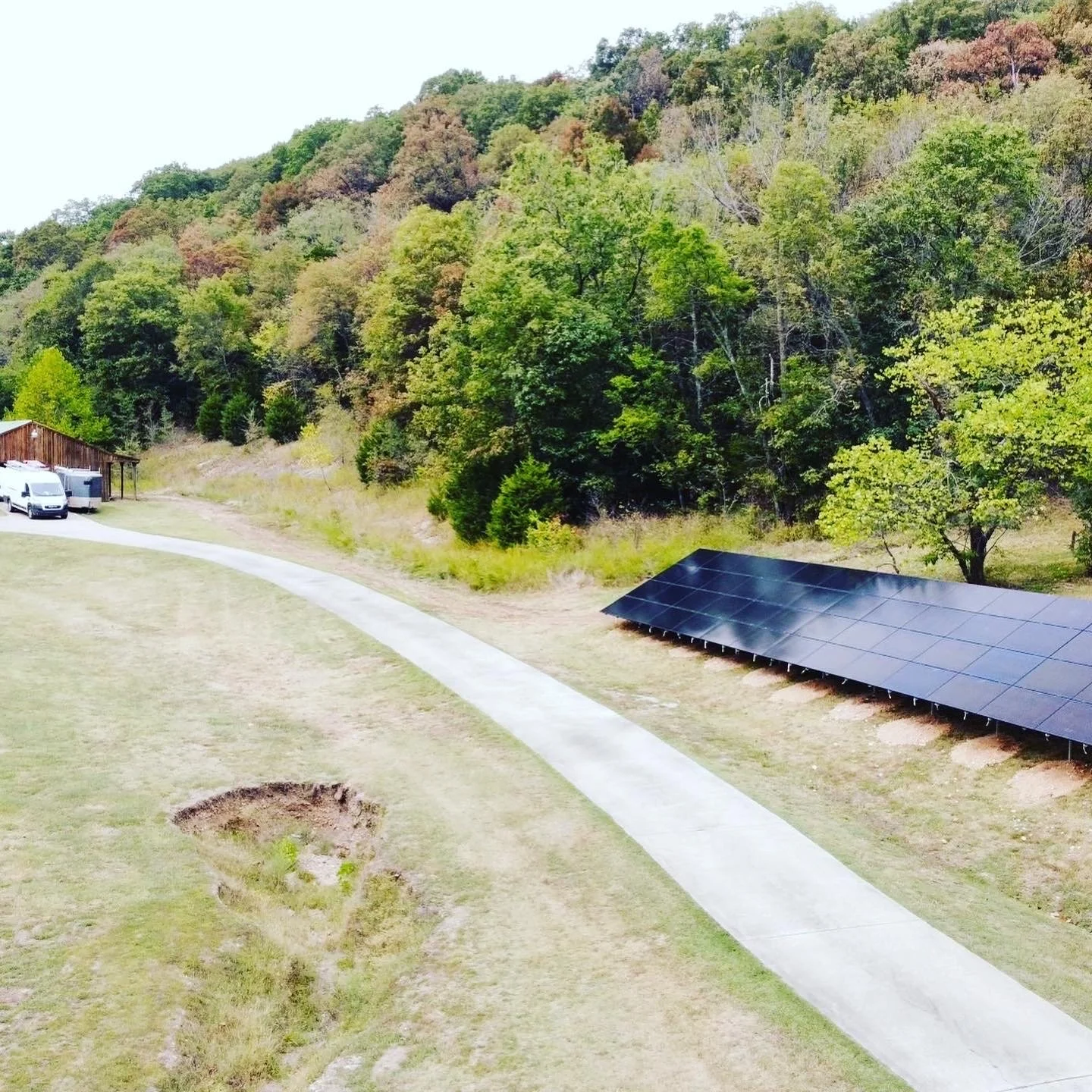Solar panels installed on the ground next to a concrete walkway and green grass, with a wooded hillside in the background.
