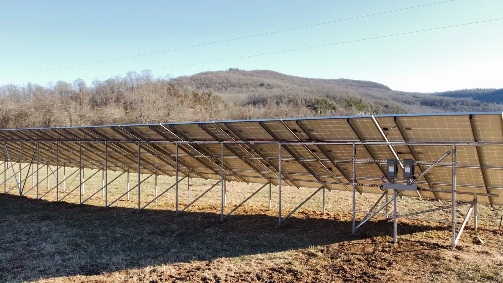Solar panel array installed on a metal scaffolding structure in an open field with hills and leafless trees in the background.