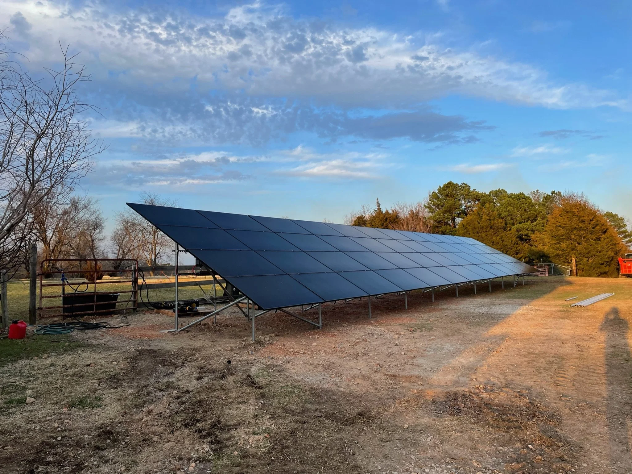 An outdoor solar panel installation on a grassy field with bare trees and some trees with green and orange leaves in the background, under a partly cloudy sky.