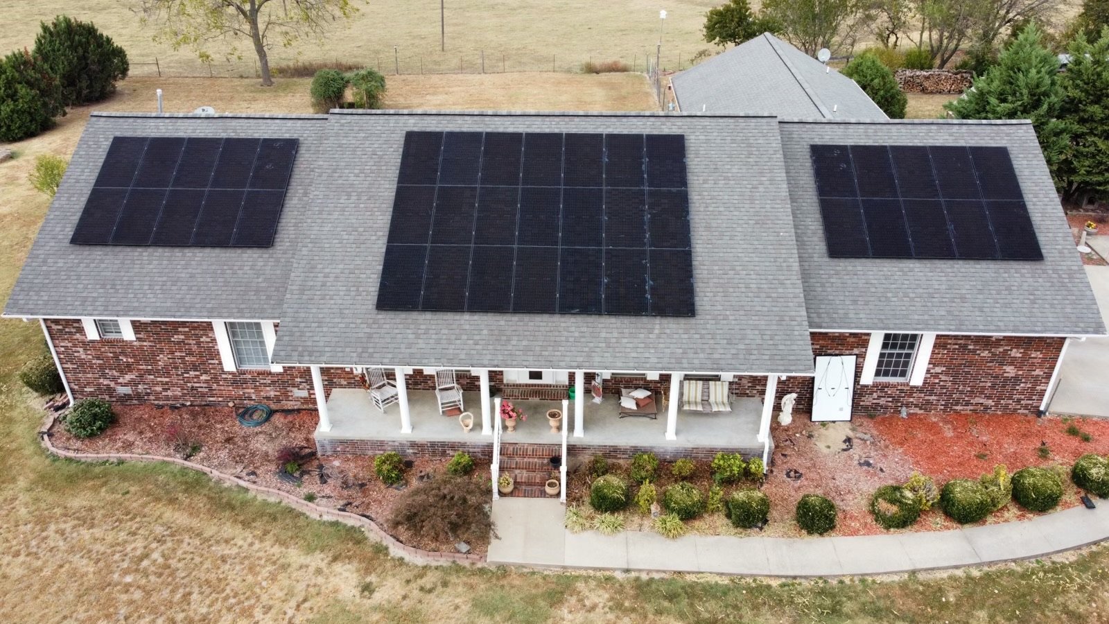 Aerial view of a brick house with a gray roof and three sets of solar panels installed on the roof.