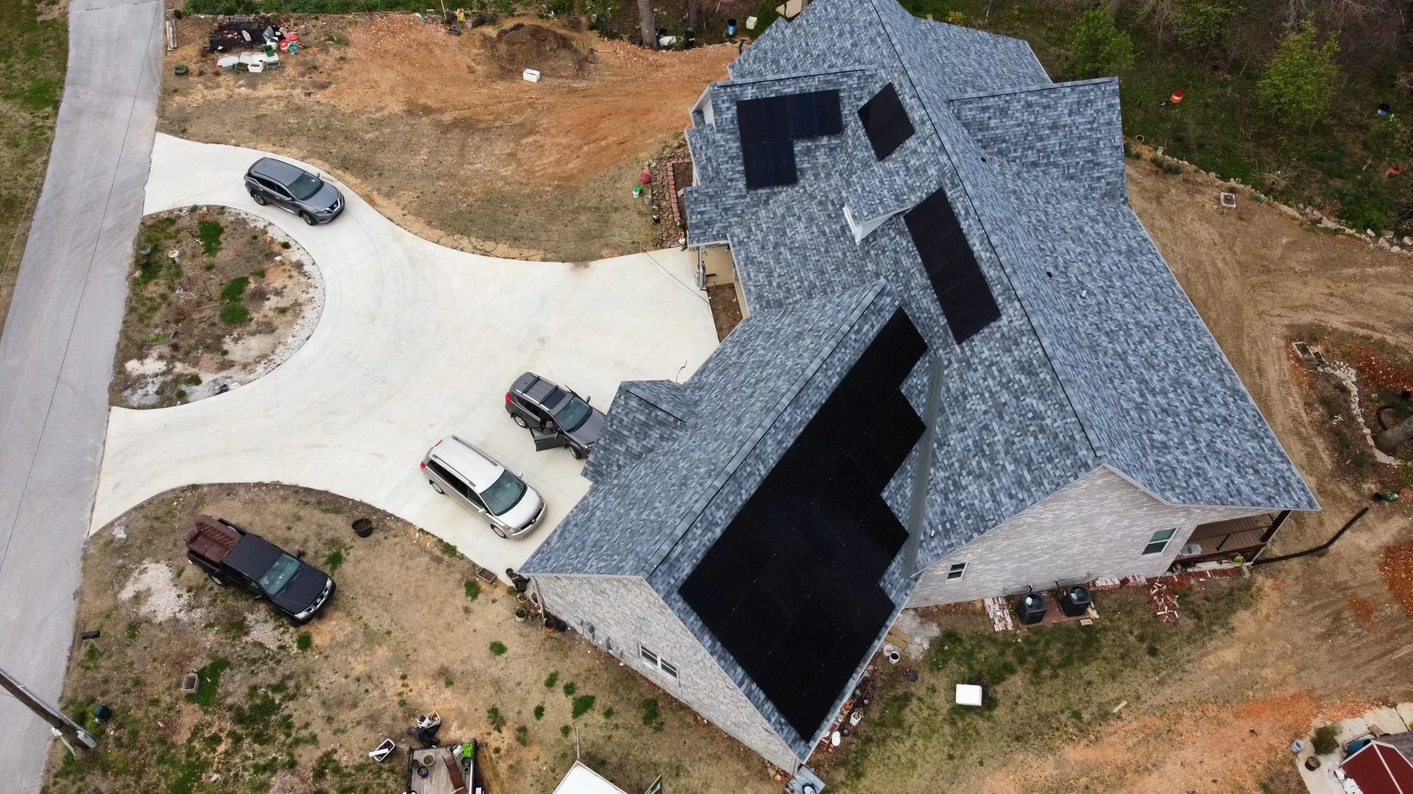 An aerial view of a residential house with solar panels on the roof, a curved concrete driveway with parked cars, and a backyard surrounded by trees and unlandscaped dirt areas.