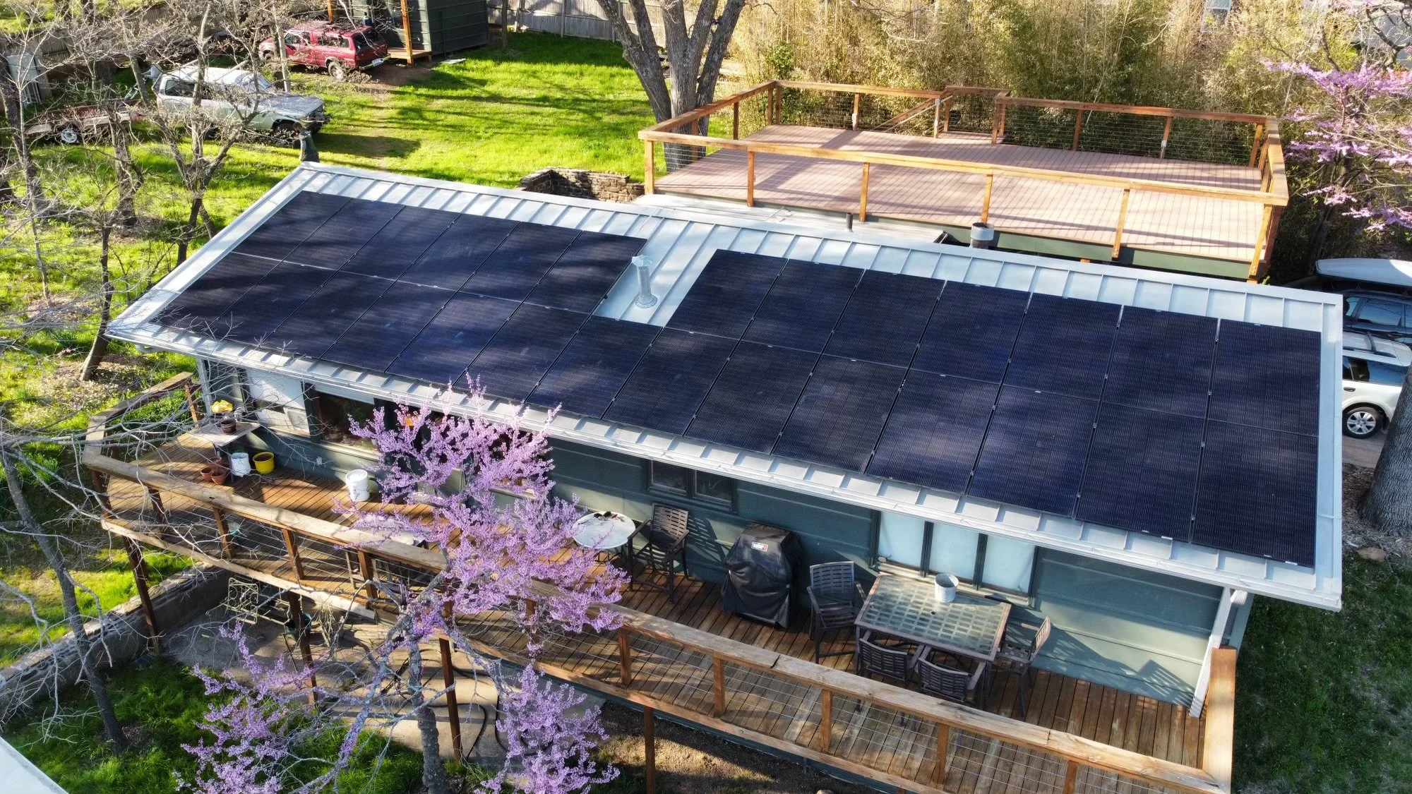 A house with a rooftop covered in solar panels and a large wooden deck, surrounded by trees including one with pink blossoms, in a green yard with a parking area.