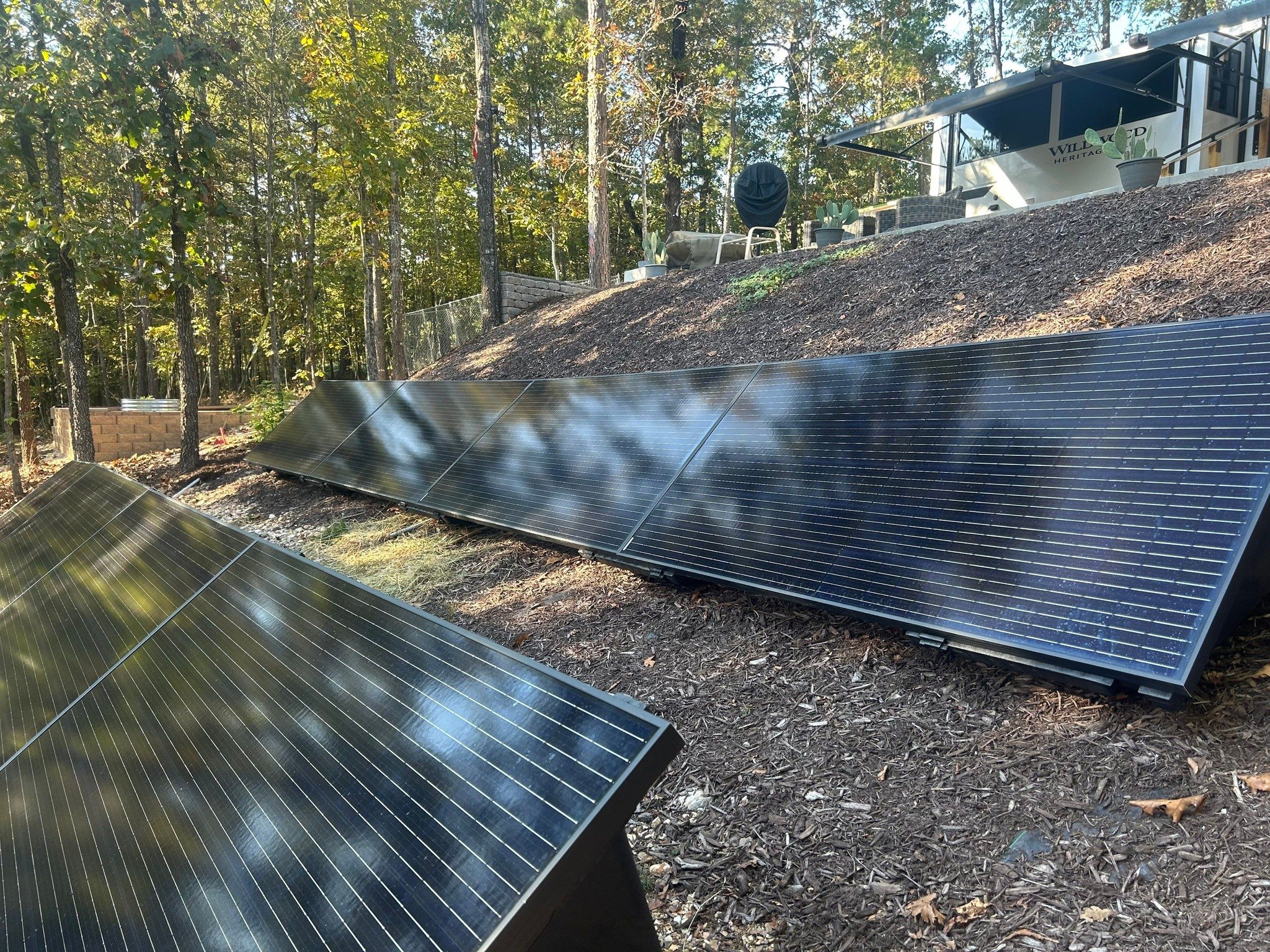 Solar panels installed on the ground in a wooded area with trees and a fence in the background.