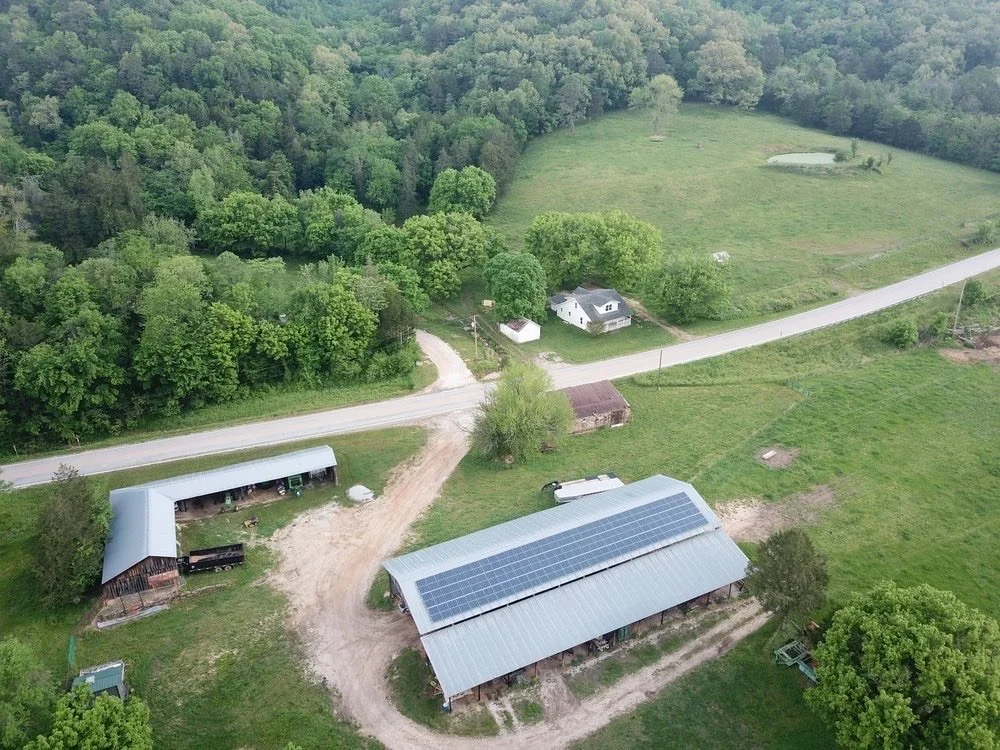 Aerial view of a rural property with farm buildings, a house with solar panels, a gravel driveway, and surrounding green trees and grassy areas.