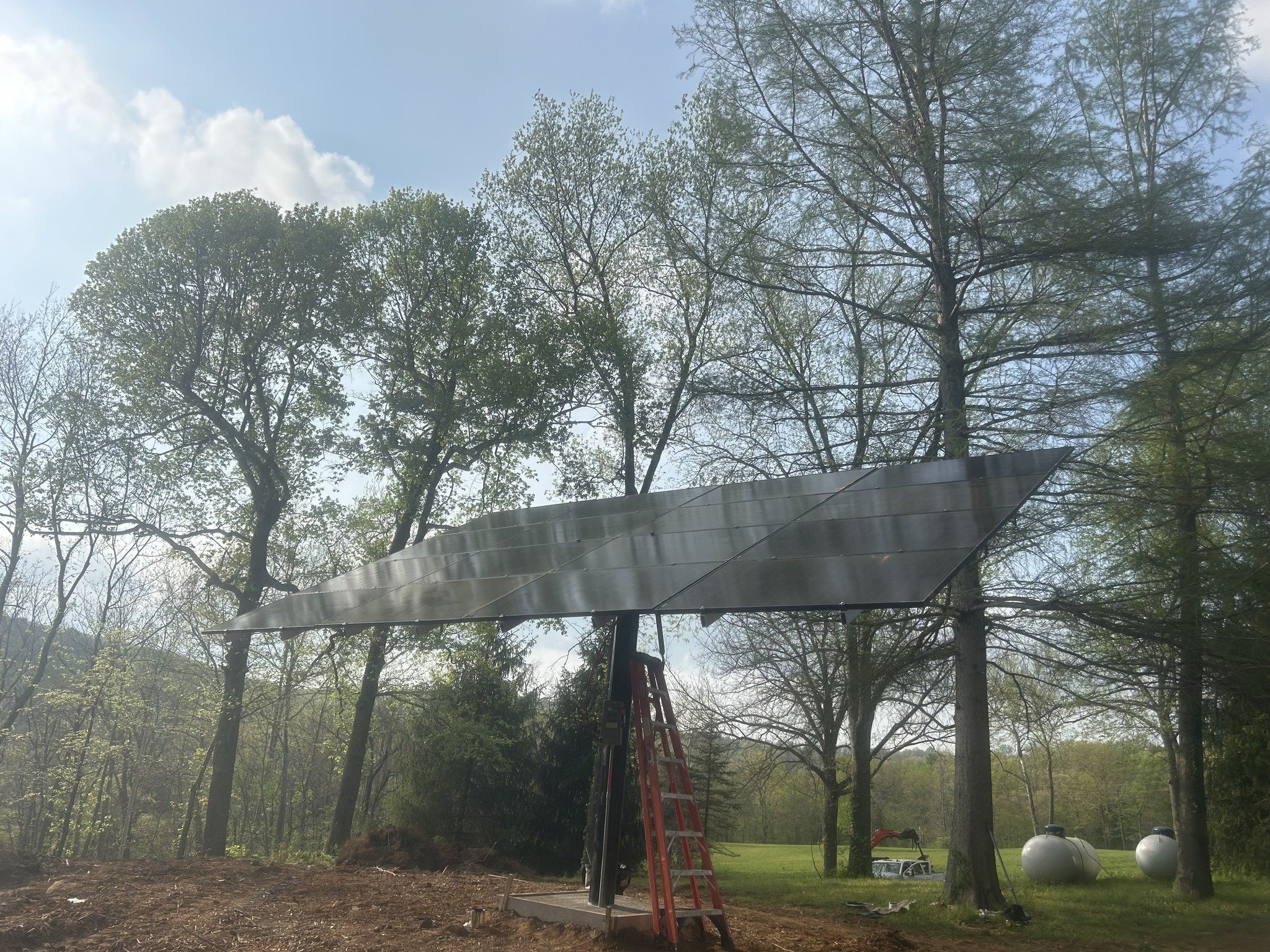 Construction work in a backyard with tall trees, a red ladder, and propane tanks, under a partly cloudy sky.