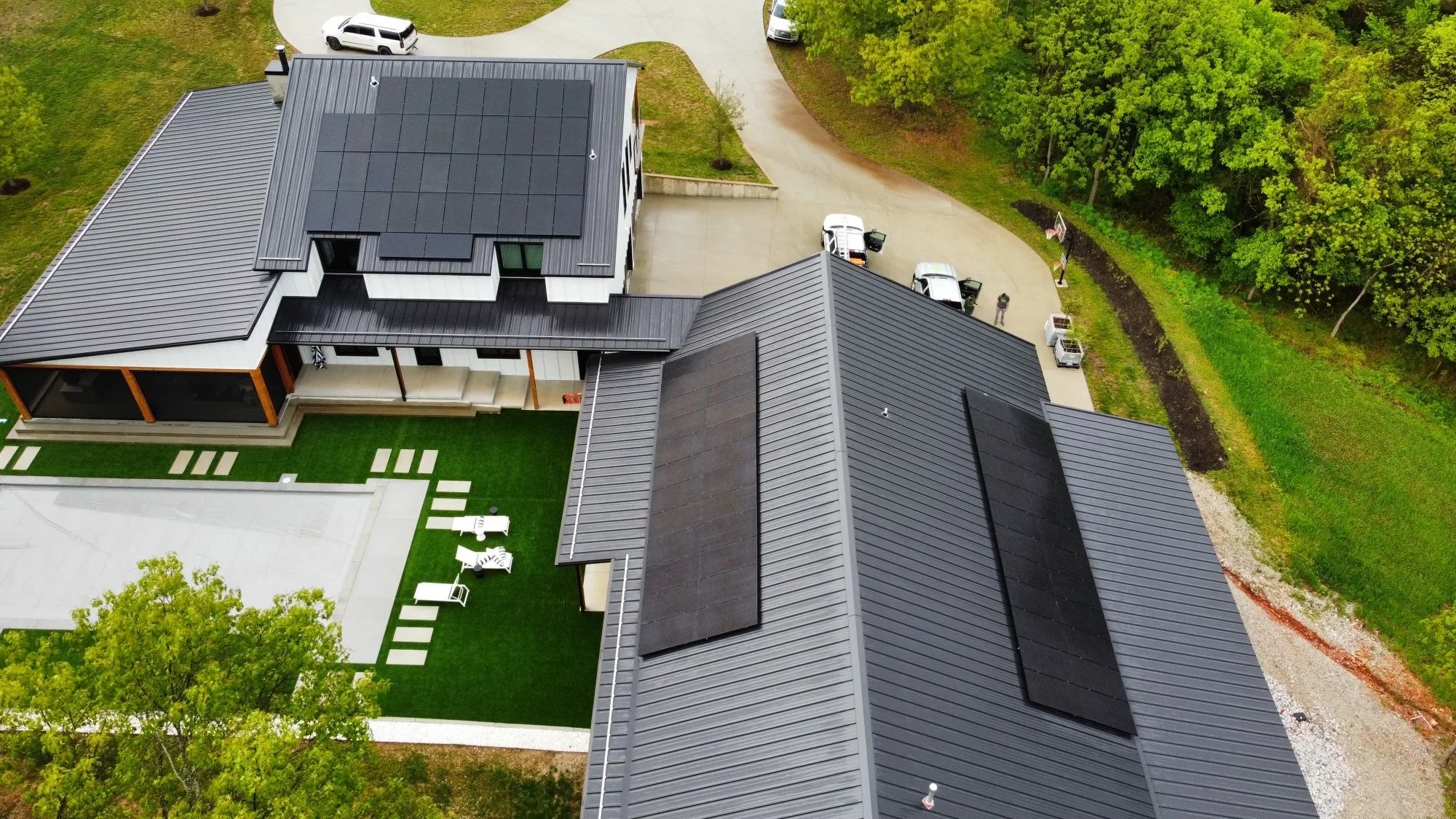 Aerial view of a modern house with solar panels on the roof, surrounded by trees and a green lawn, with a driveway and parked cars.