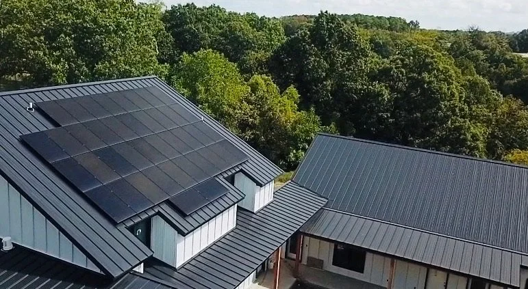 Aerial view of rooftops with solar panels on one roof, surrounded by green trees in the background.