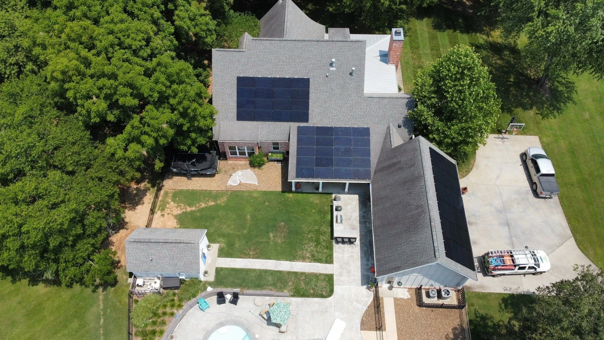 Aerial view of a house with solar panels on the roof, surrounded by trees, a driveway with parked vehicle, a small pool, and a backyard area with outdoor furniture.