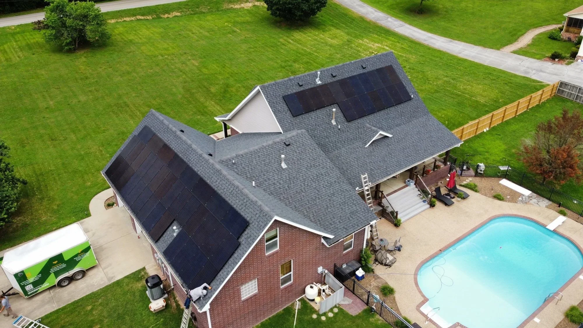 Aerial view of a house with solar panels on the roof, a backyard with a swimming pool, patio furniture, trees, and adjacent green lawn areas.