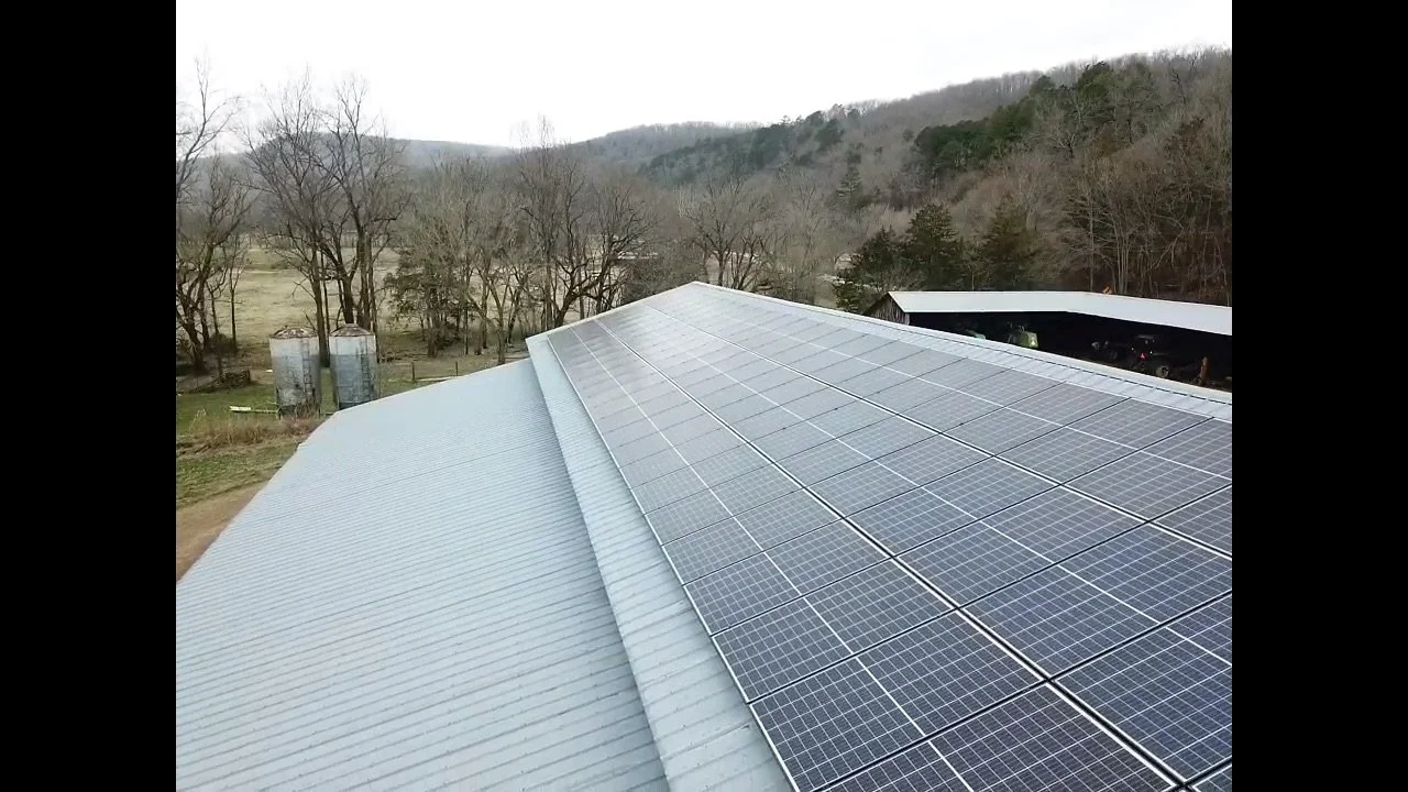 Solar panels installed on the roof of a building in a rural area with trees and mountains in the background.