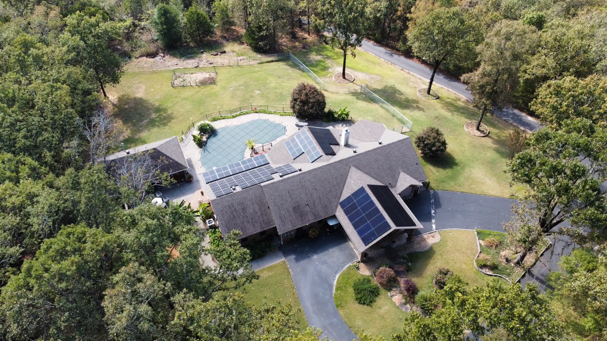 Aerial view of a house with solar panels on the roof, a backyard with a screened swimming pool, trees, and a driveway on a sunny day.