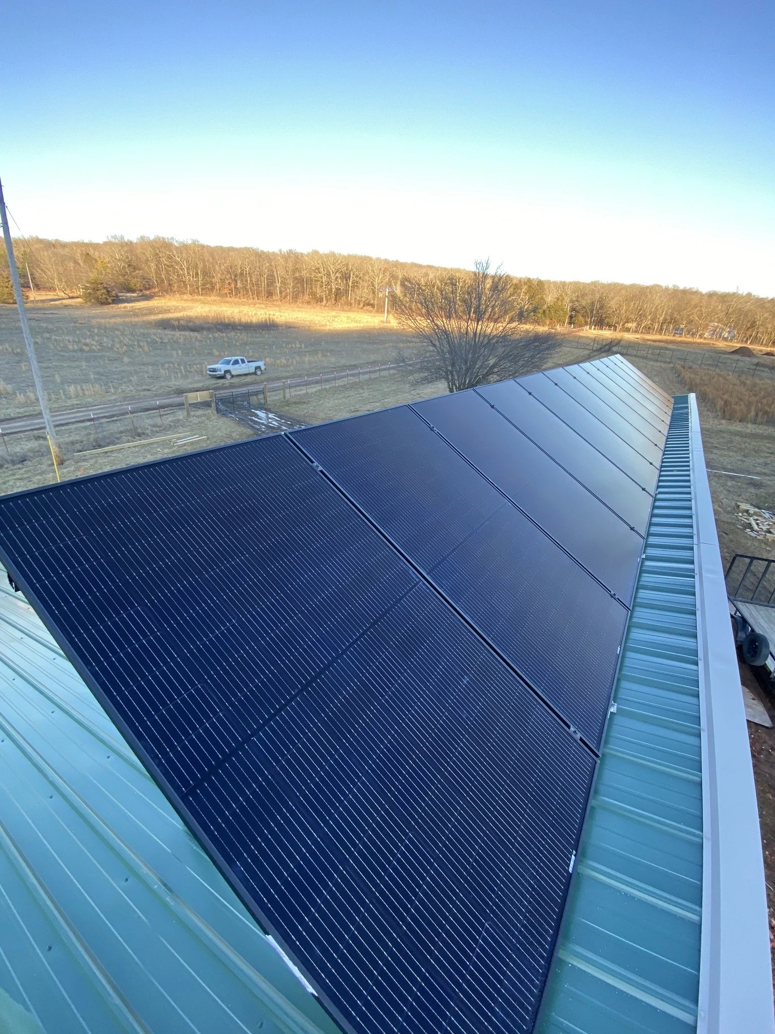 View of solar panels installed on a roof with a rural landscape in the background, including a field, trees, and a pickup truck.