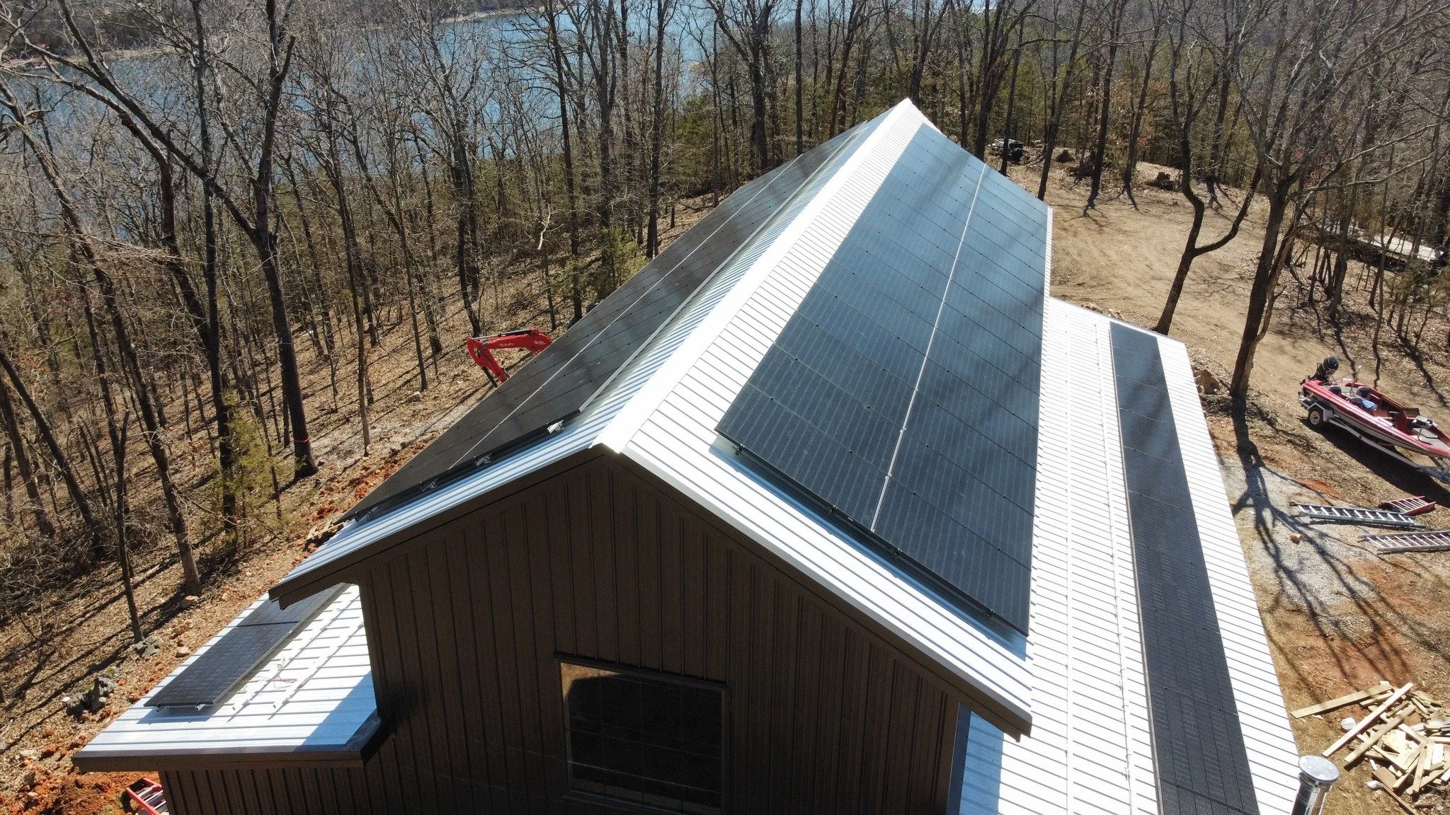 Aerial view of a house with solar panels on the roof in a wooded area with a lake nearby, construction equipment and a boat on the ground.