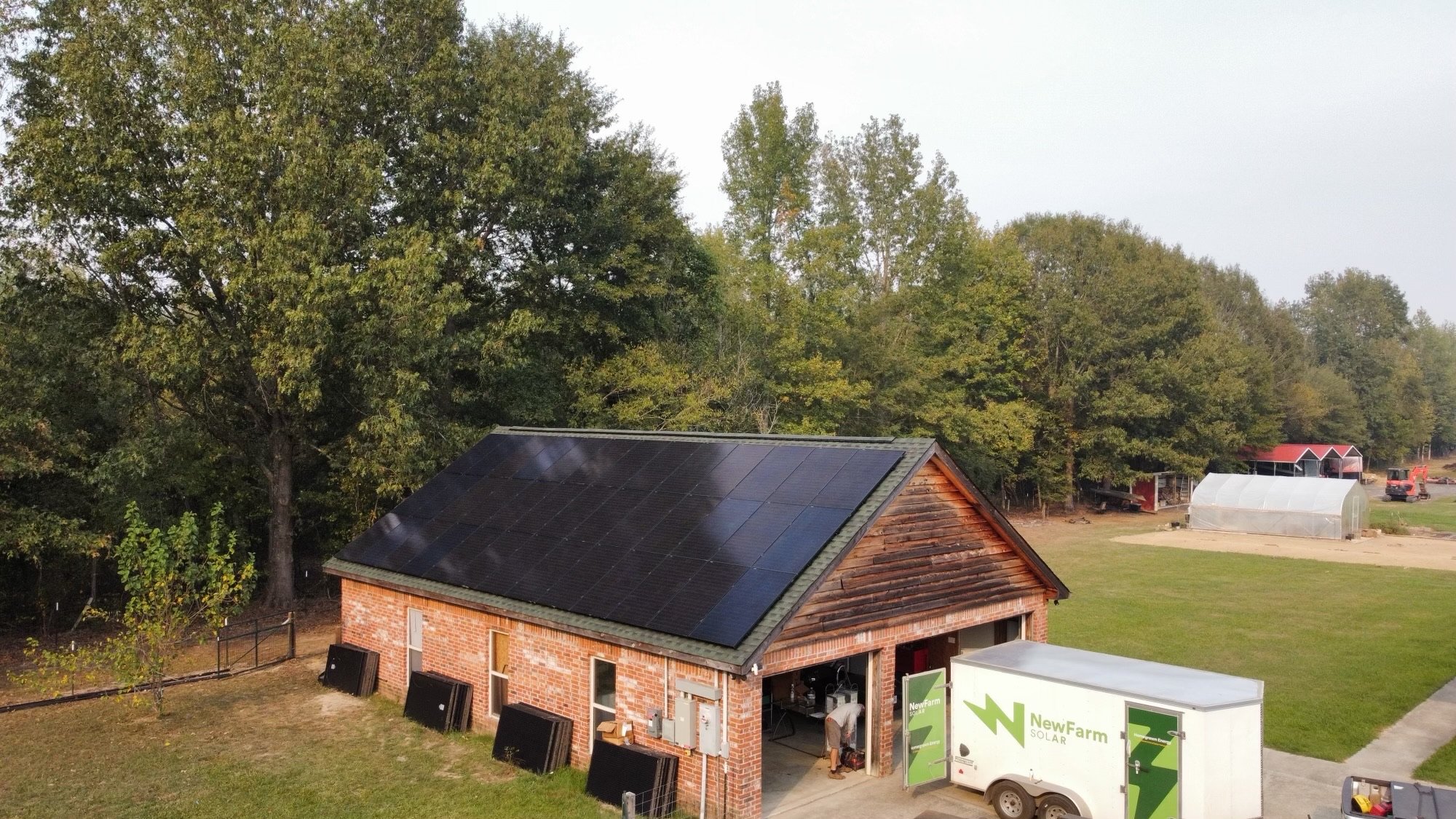 A brick building with a black solar panel roof in a rural area surrounded by trees and other farm structures, including a greenhouse and a red barn.