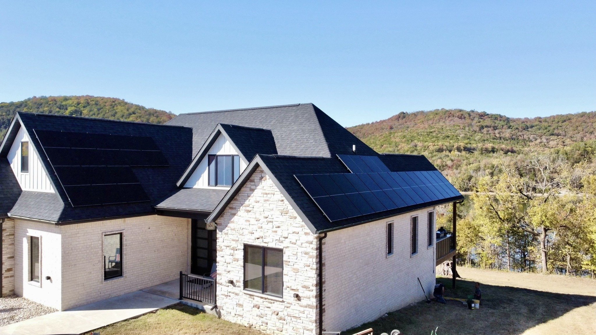 A house with a black roof and solar panels, set against a backdrop of green mountains and a clear blue sky.