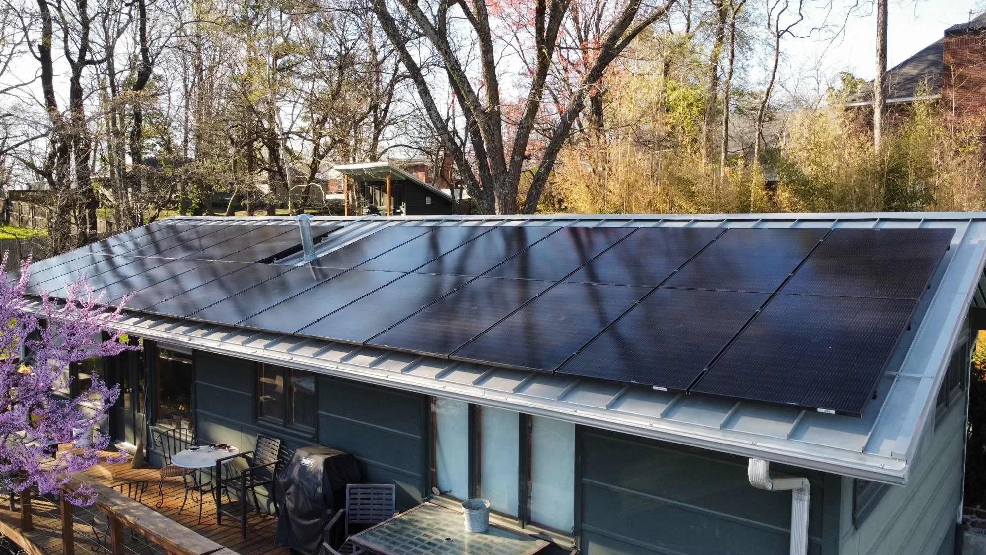 House rooftop with solar panels, surrounded by trees and backyard patio furniture.