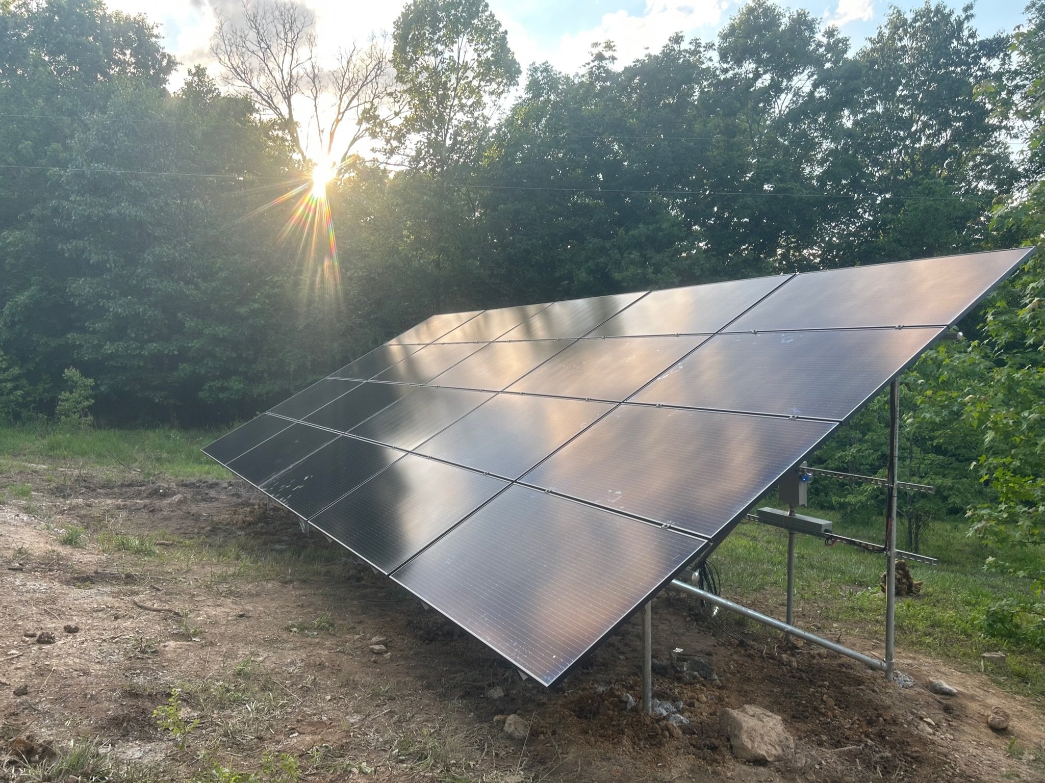 Solar panels installed on the ground in a grassy area with trees and the sun shining through the leaves.