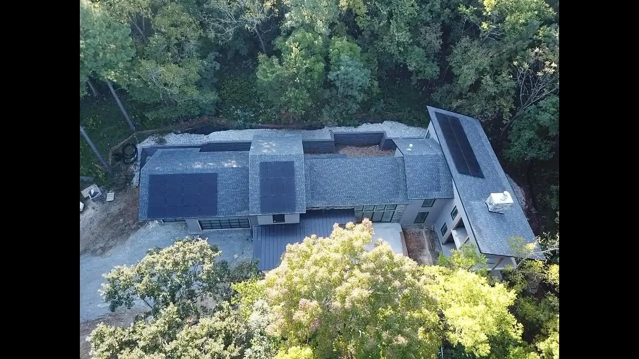 Aerial view of a modern house surrounded by trees, with solar panels on the roof and a gravel driveway.