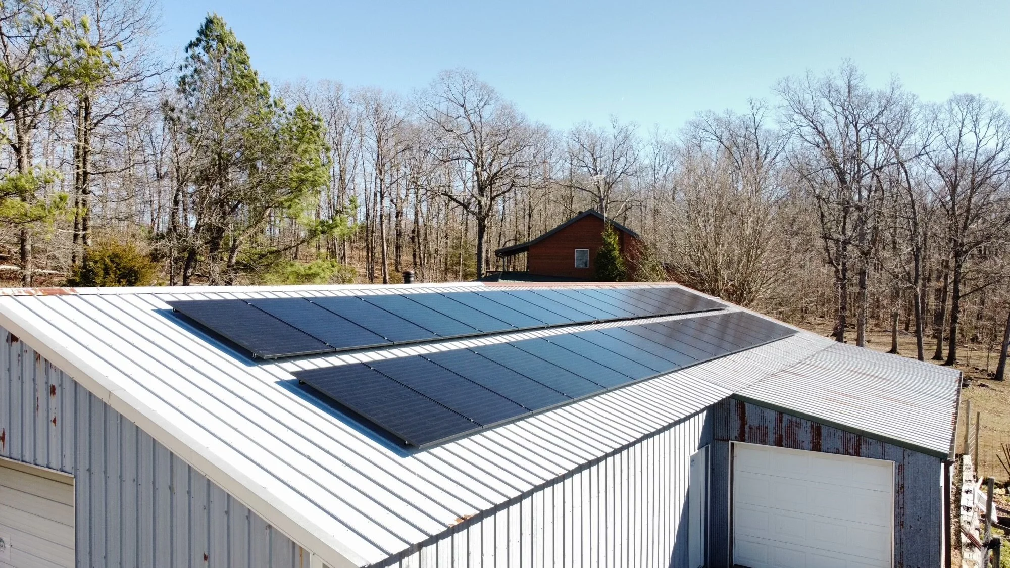 Metal building with solar panels on the roof, surrounded by trees and a brown house in the background during daytime.