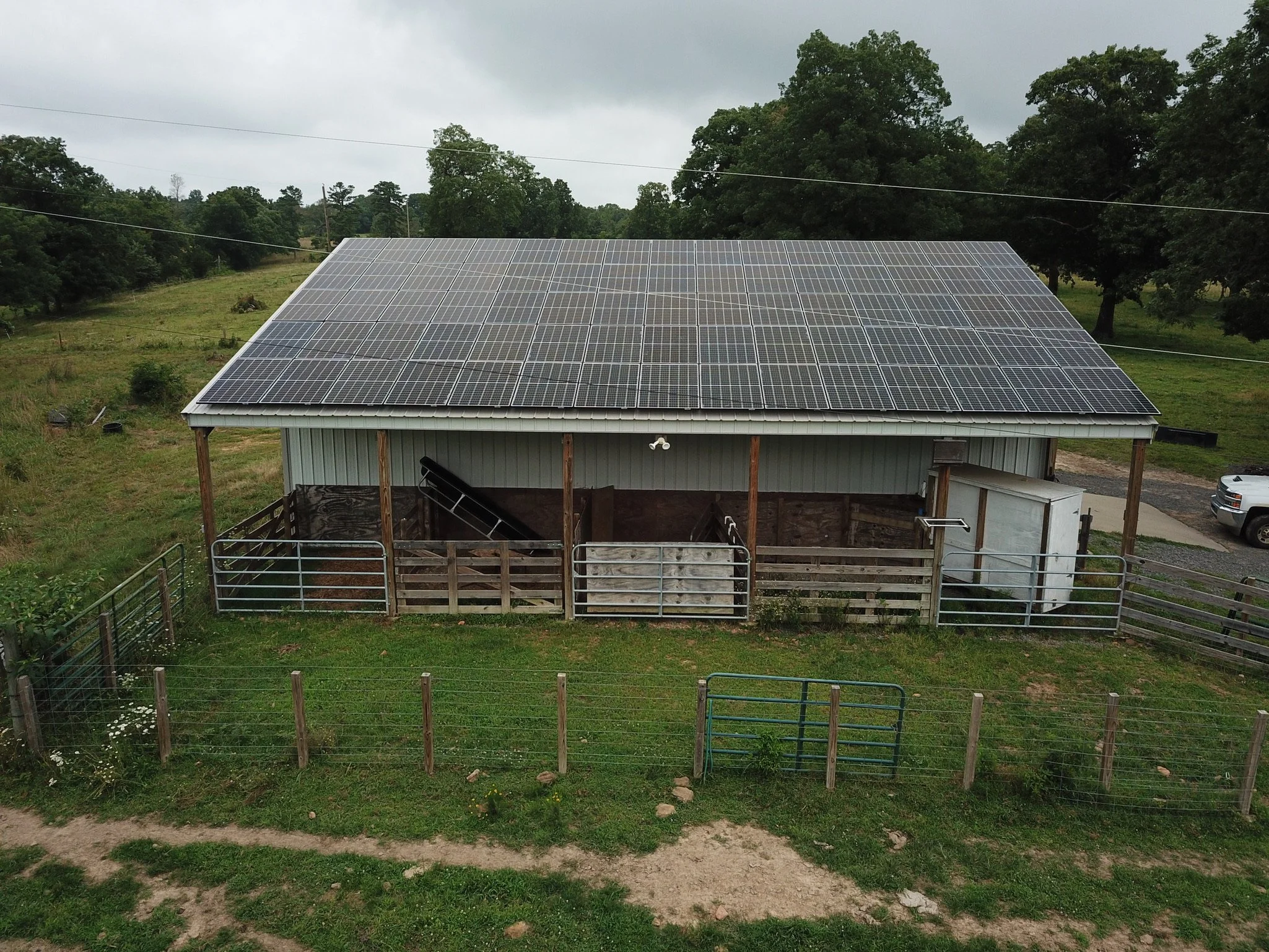 A house with solar panels on the roof, surrounded by trees and a fenced yard, on a cloudy day.