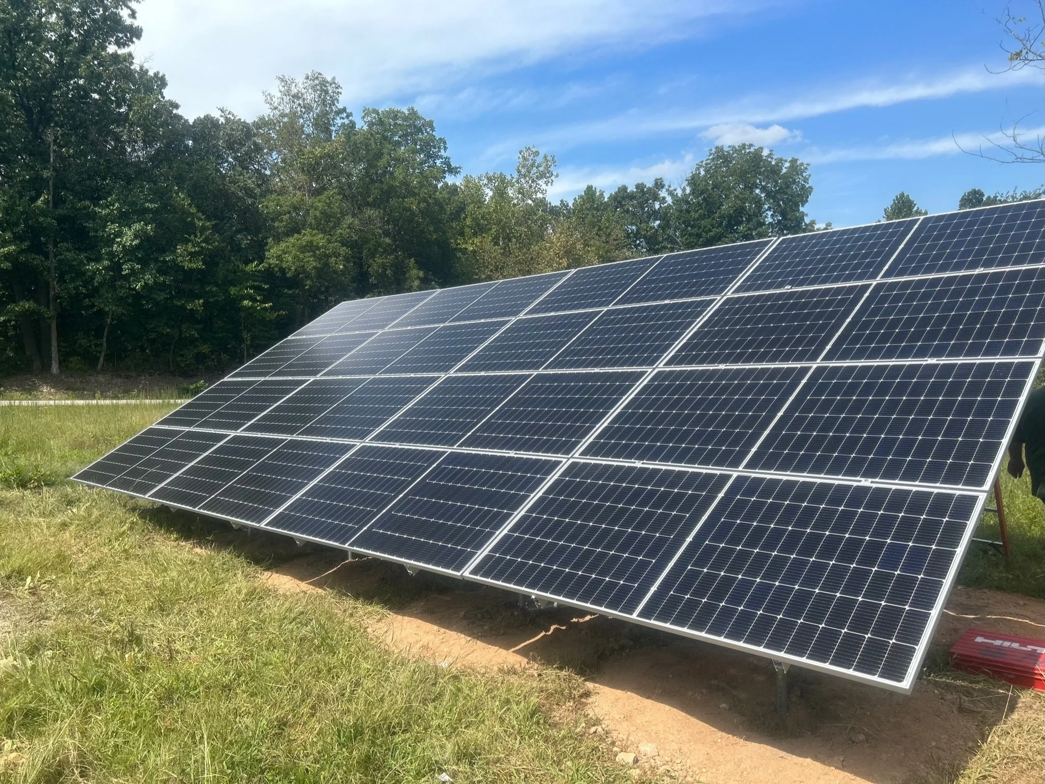 Multiple solar panels installed on grassy ground with trees and blue sky in the background.