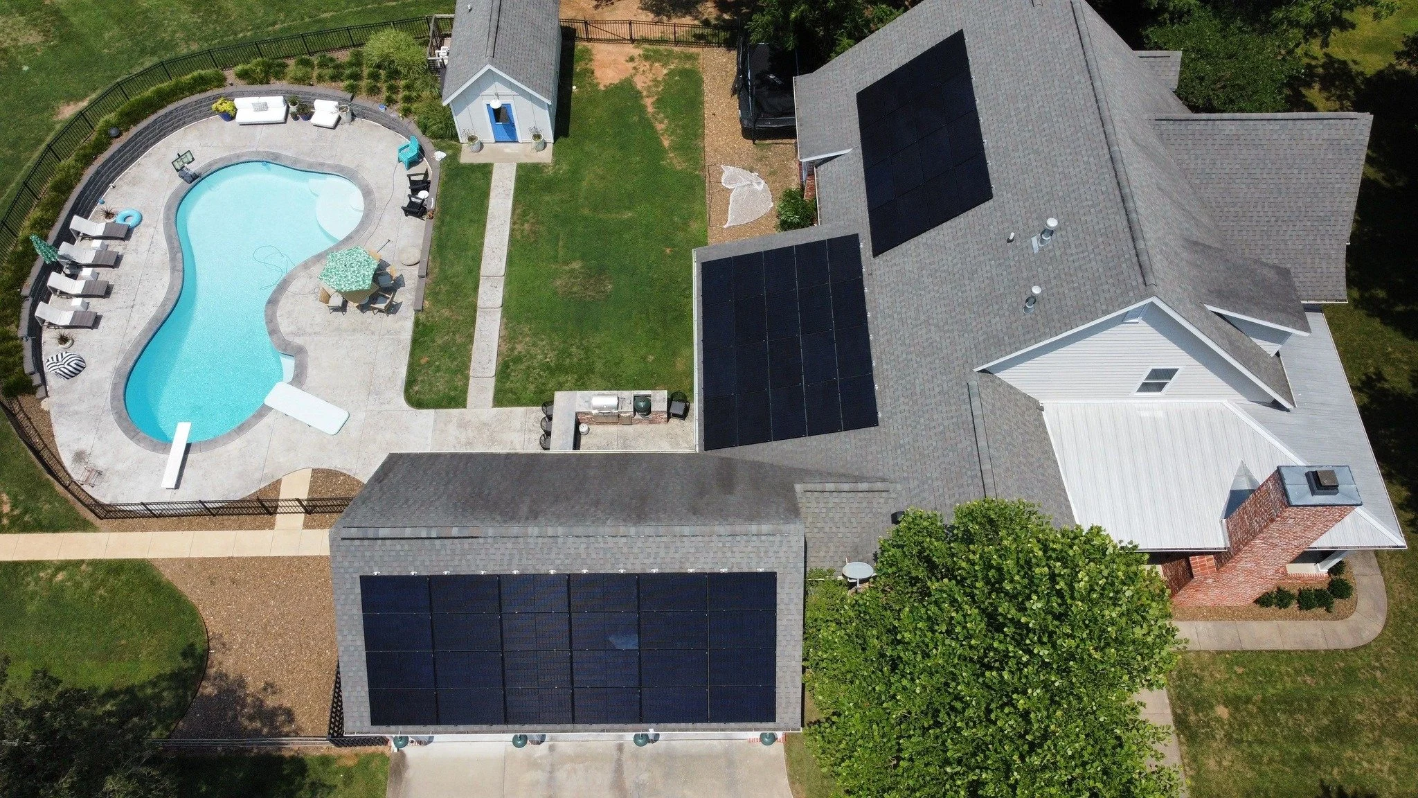 Aerial view of a residential backyard showing a swimming pool with surrounding patio furniture, a small white shed, a grassy area, and a house with solar panels on the roof.