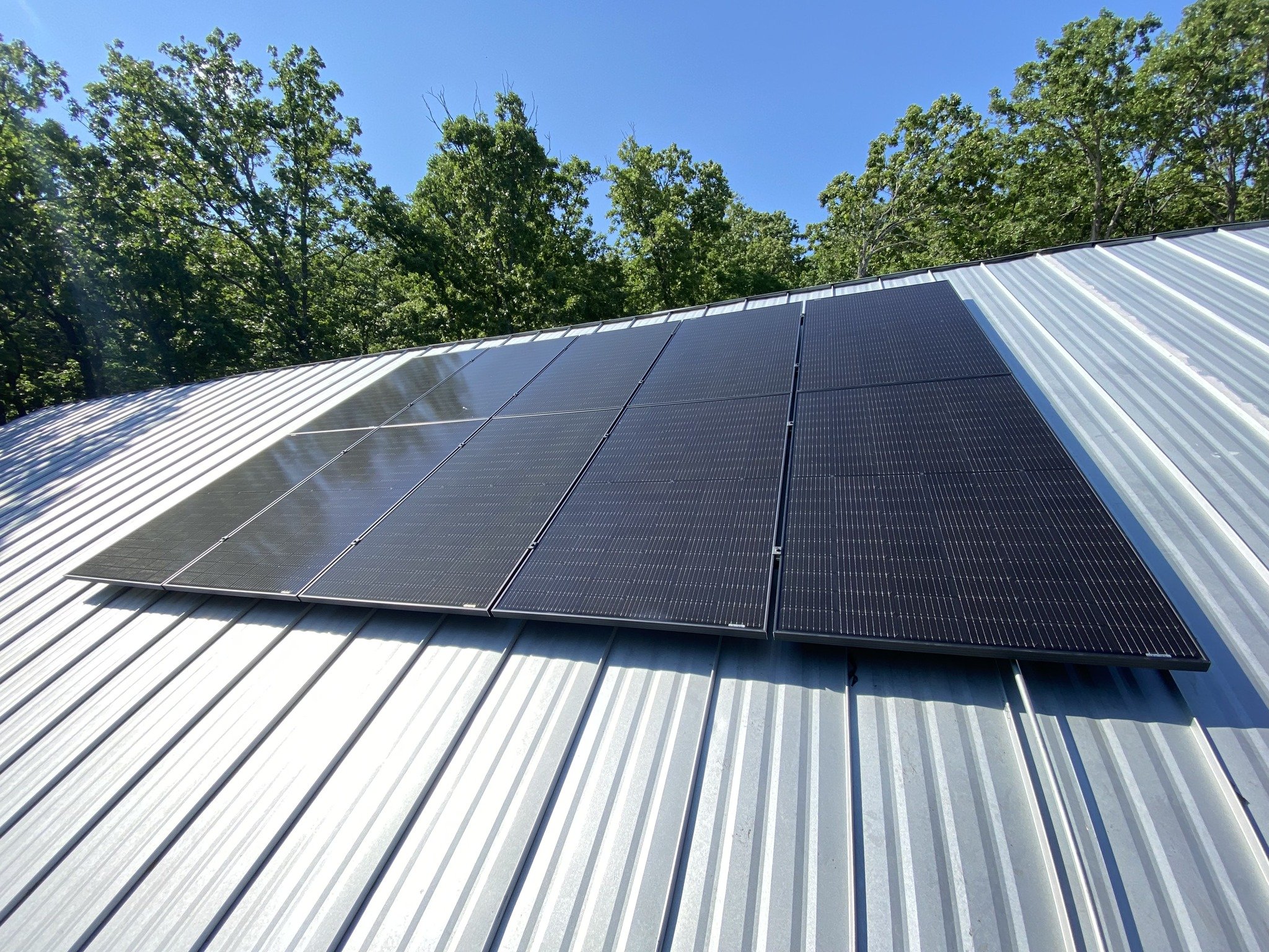 Solar panels installed on a metal roof with a background of green trees and a clear blue sky.