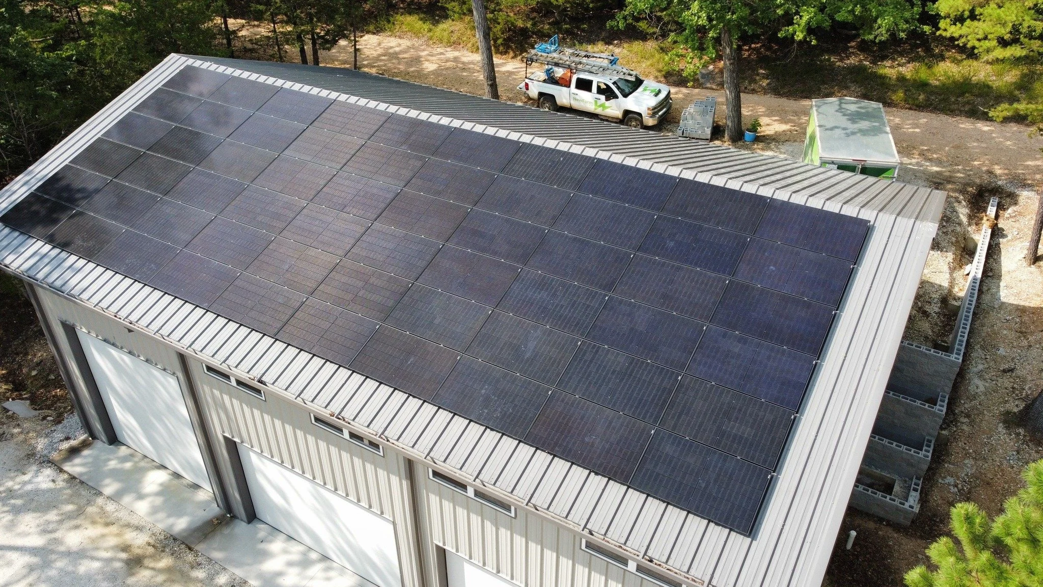 A building with a roof covered in solar panels, surrounded by trees and outdoor equipment, including a white utility truck and a small covered structure.