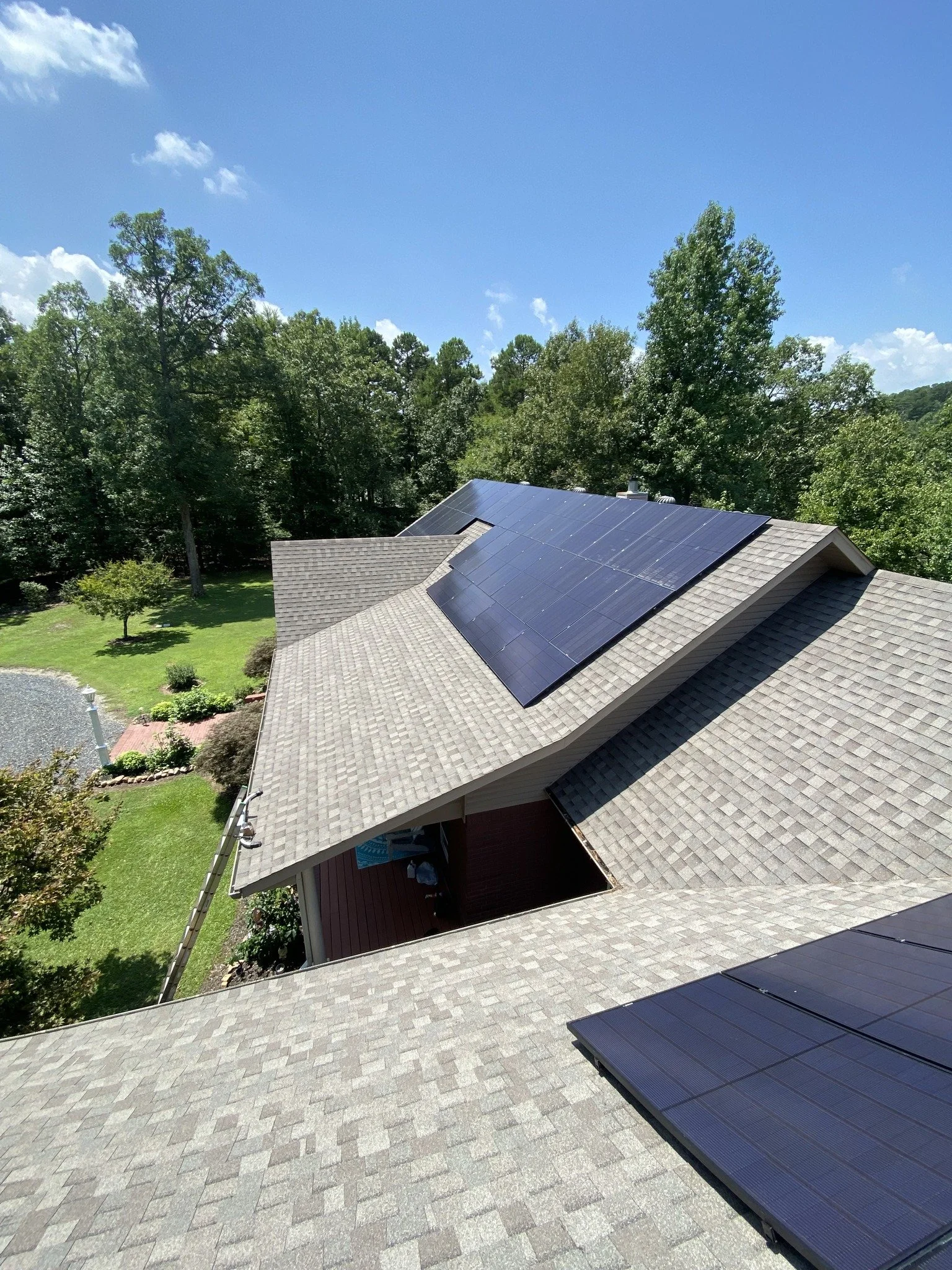 Residential house with solar panels on the roof, surrounded by green trees and a well-maintained yard, under a bright blue sky with some clouds.