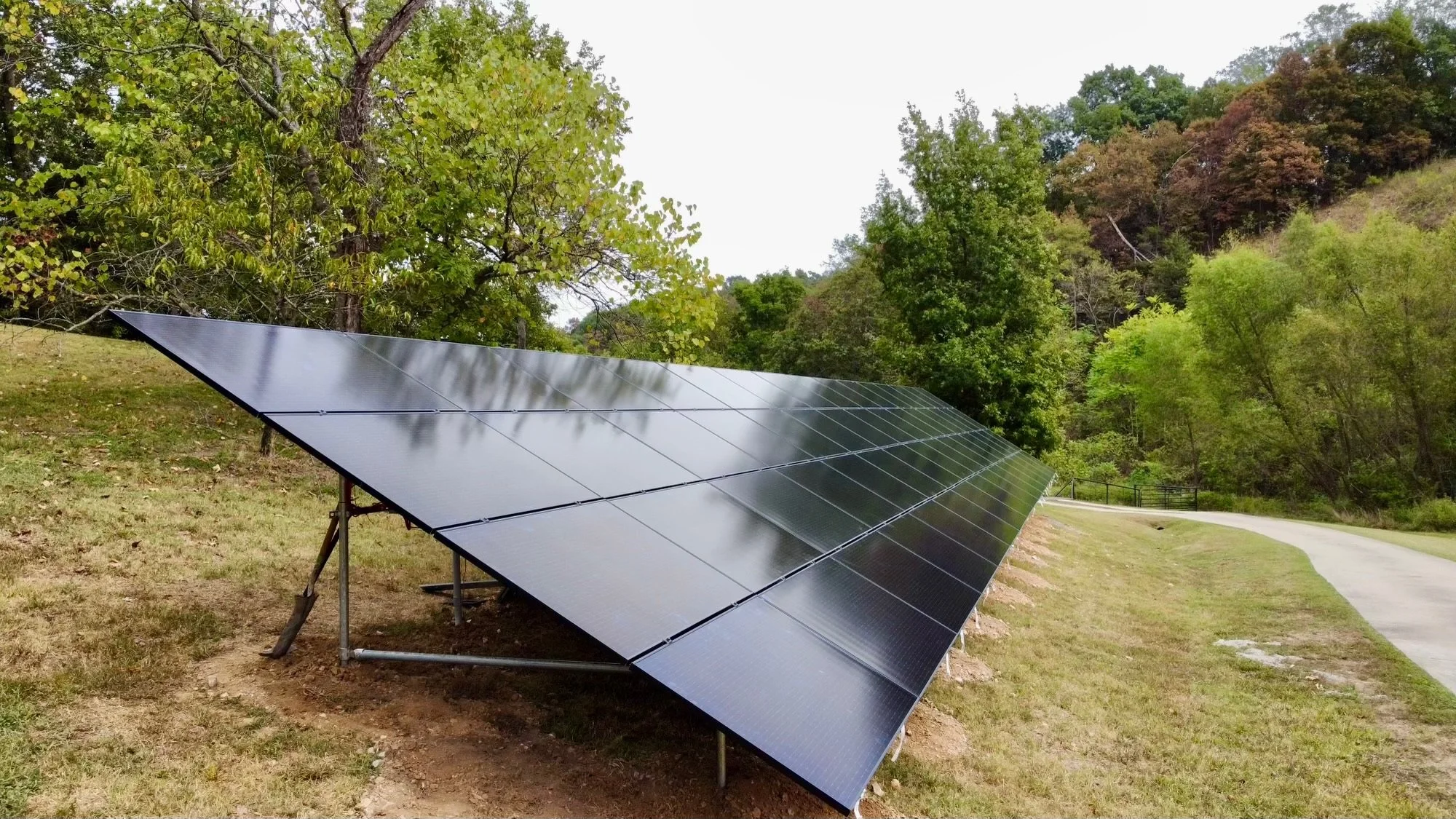 Solar panels installed on a grassy hill next to a paved pathway, with green trees and a forested hillside in the background.