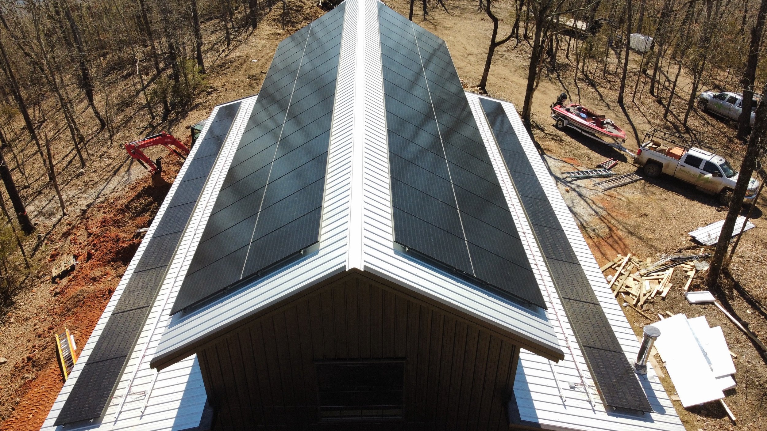 Aerial view of a building with black solar panels on its white metal roof, surrounded by trees and construction equipment.