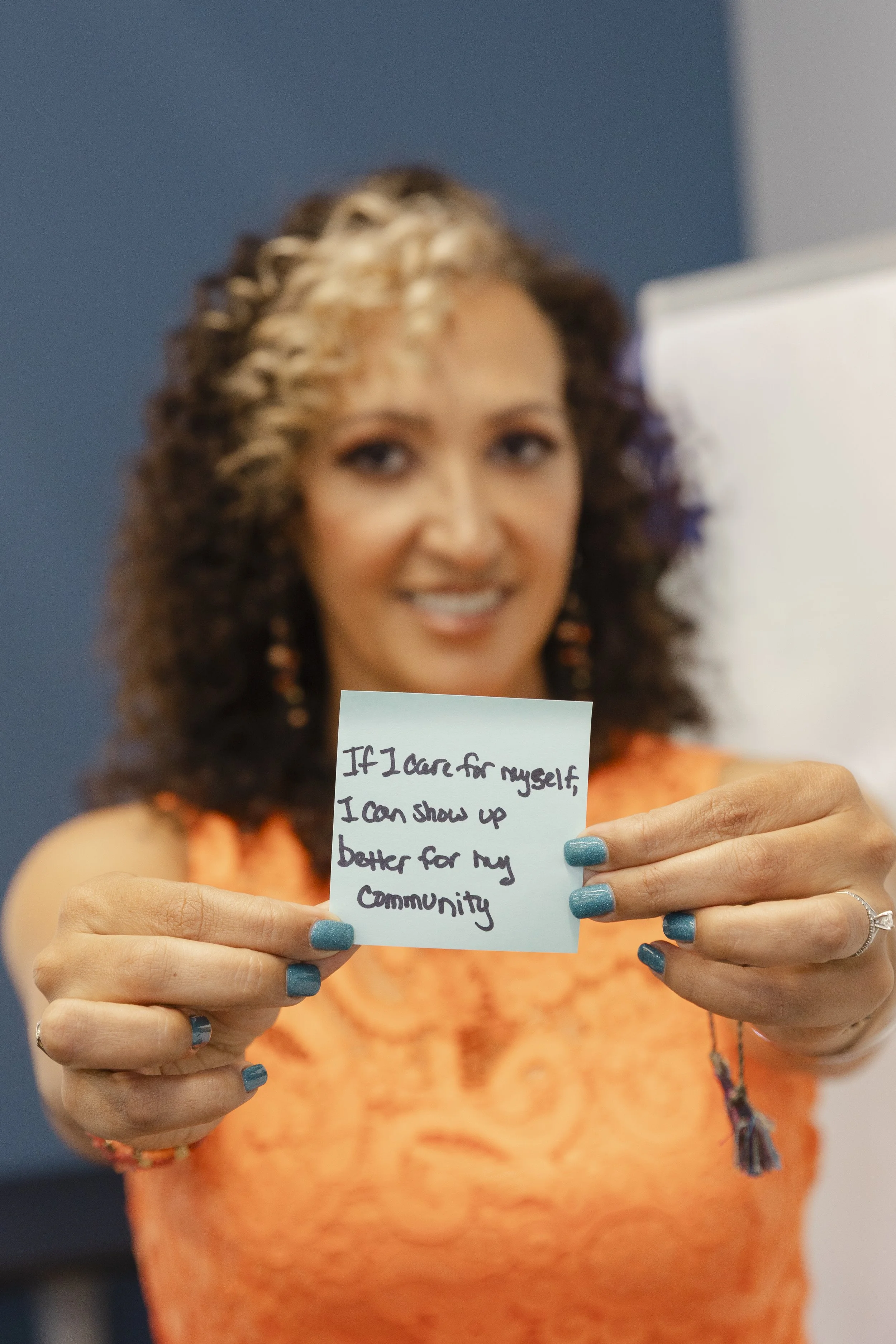 A woman with curly hair holding a small note that says, 'If I care for myself, I can show up better for my community'.