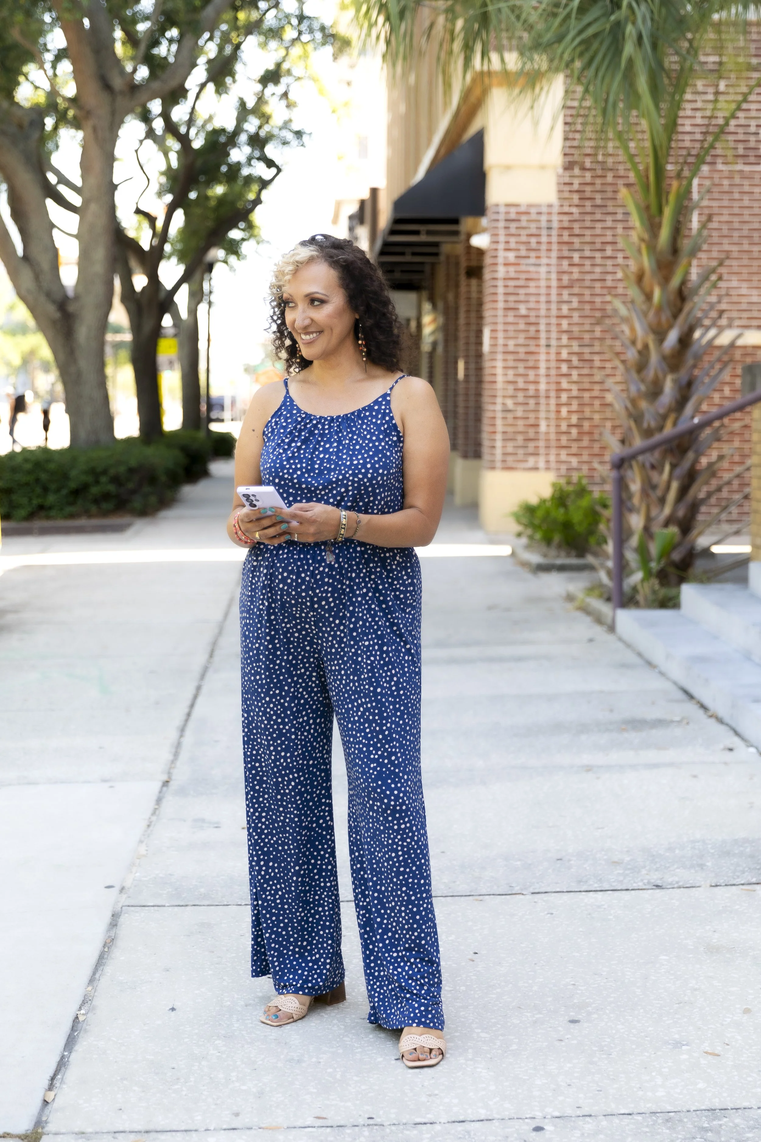 A woman with curly hair, wearing a blue polka dot jumpsuit, standing outdoors on a sidewalk with trees and buildings in the background, holding a phone and smiling.