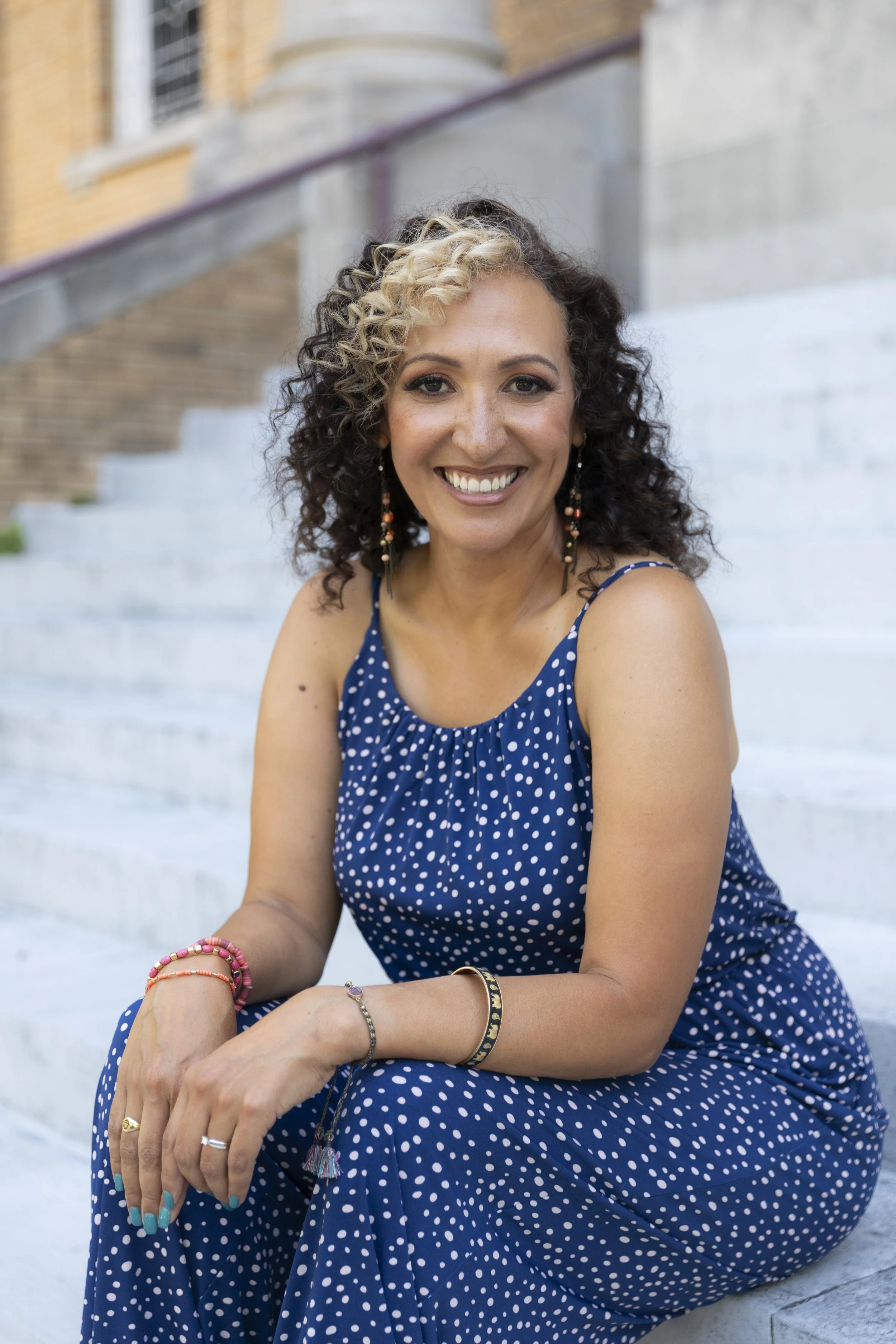 A woman with curly hair, wearing a blue polka dot dress, sitting on outdoor stairs, smiling, with colorful jewelry.