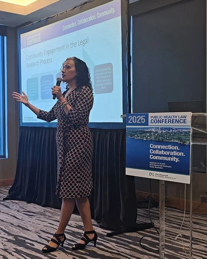 A woman in a black and pink patterned dress giving a presentation at a conference, standing next to a podium with a sign for the 2025 Public Health Law Conference, with a projection screen behind her showing slides on community engagement in legal research.