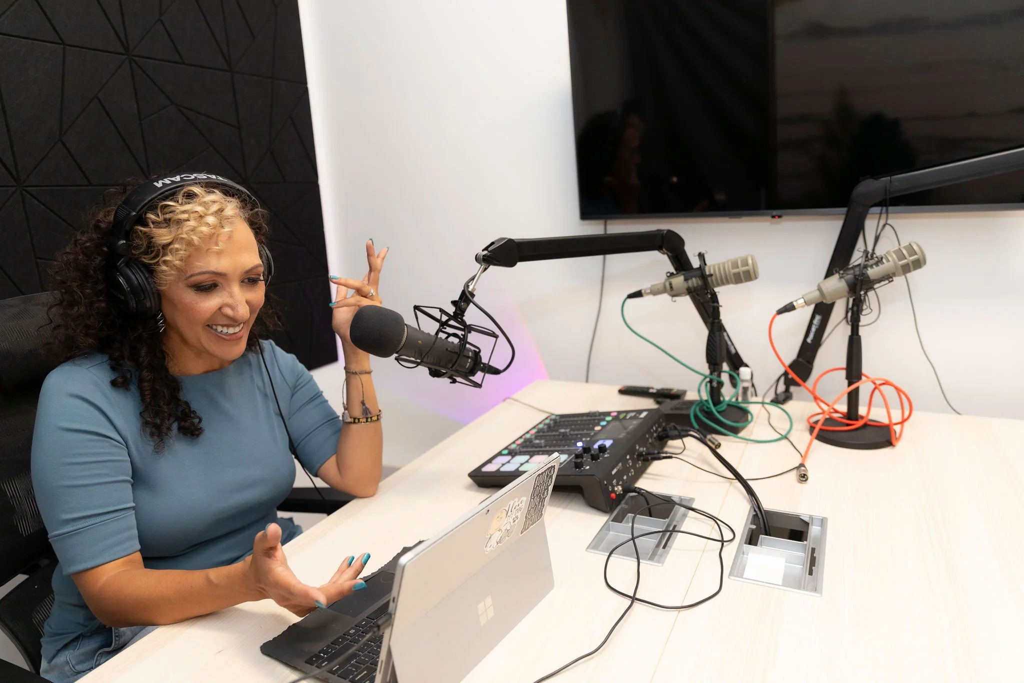 A woman with curly blonde and brown hair wearing headphones, sitting at a radio studio table with a microphone, laptop, and audio equipment, speaking and gesturing with her hands, in front of a large monitor.