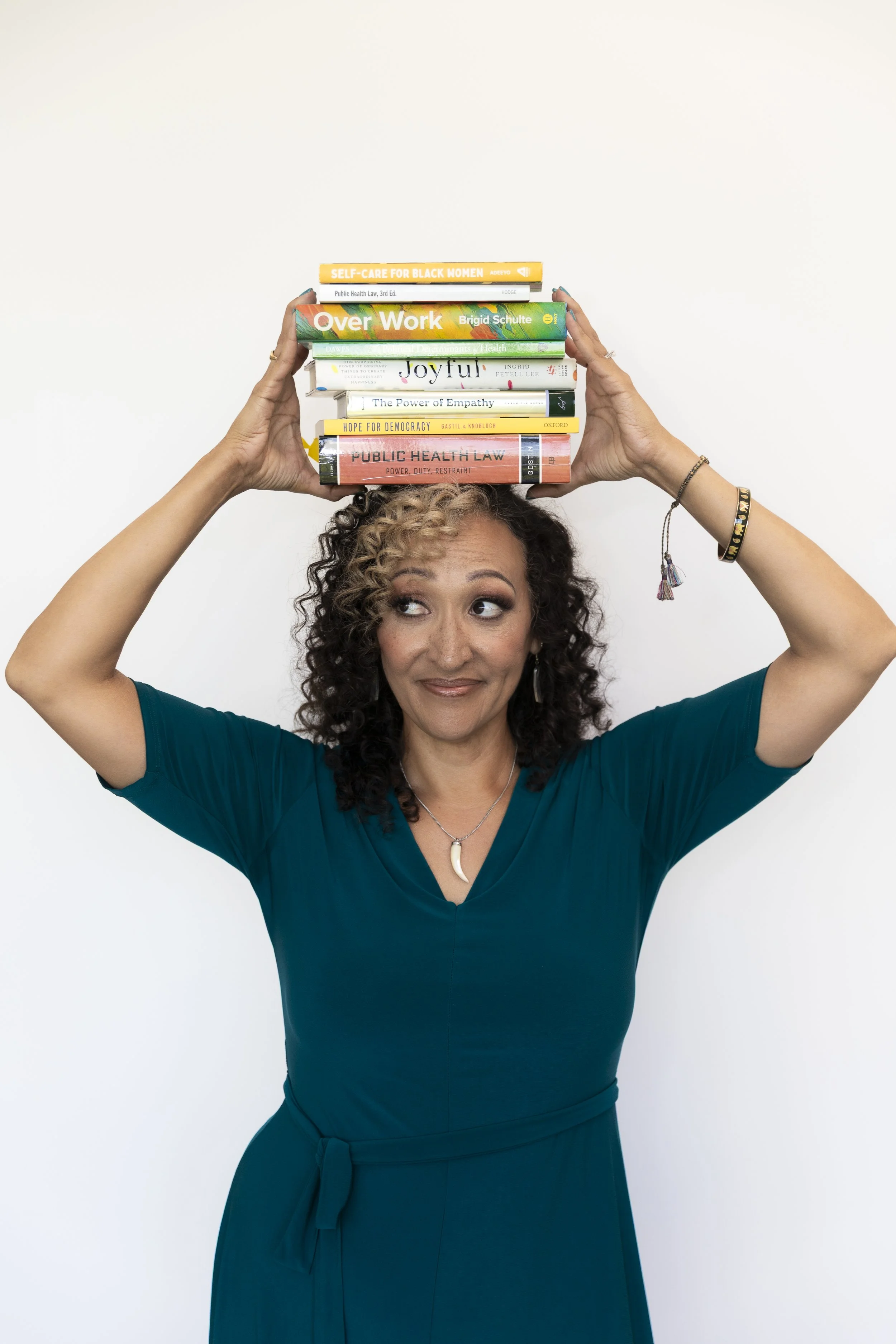 A woman with curly hair wearing a teal dress holds a stack of books on her head, looking upwards with a smile.