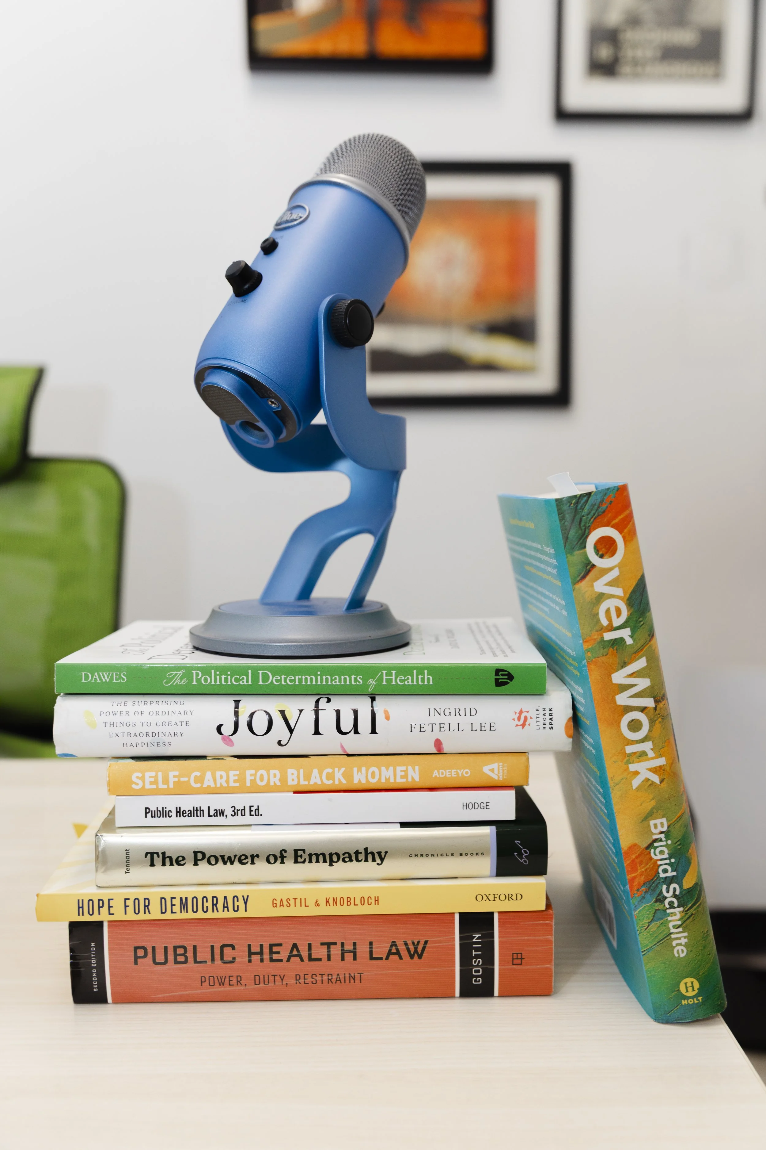 A blue microphone on a stand placed on top of several books related to health, policy, and empowerment, with a colorful stack of books to the right and framed artwork on the wall in the background.