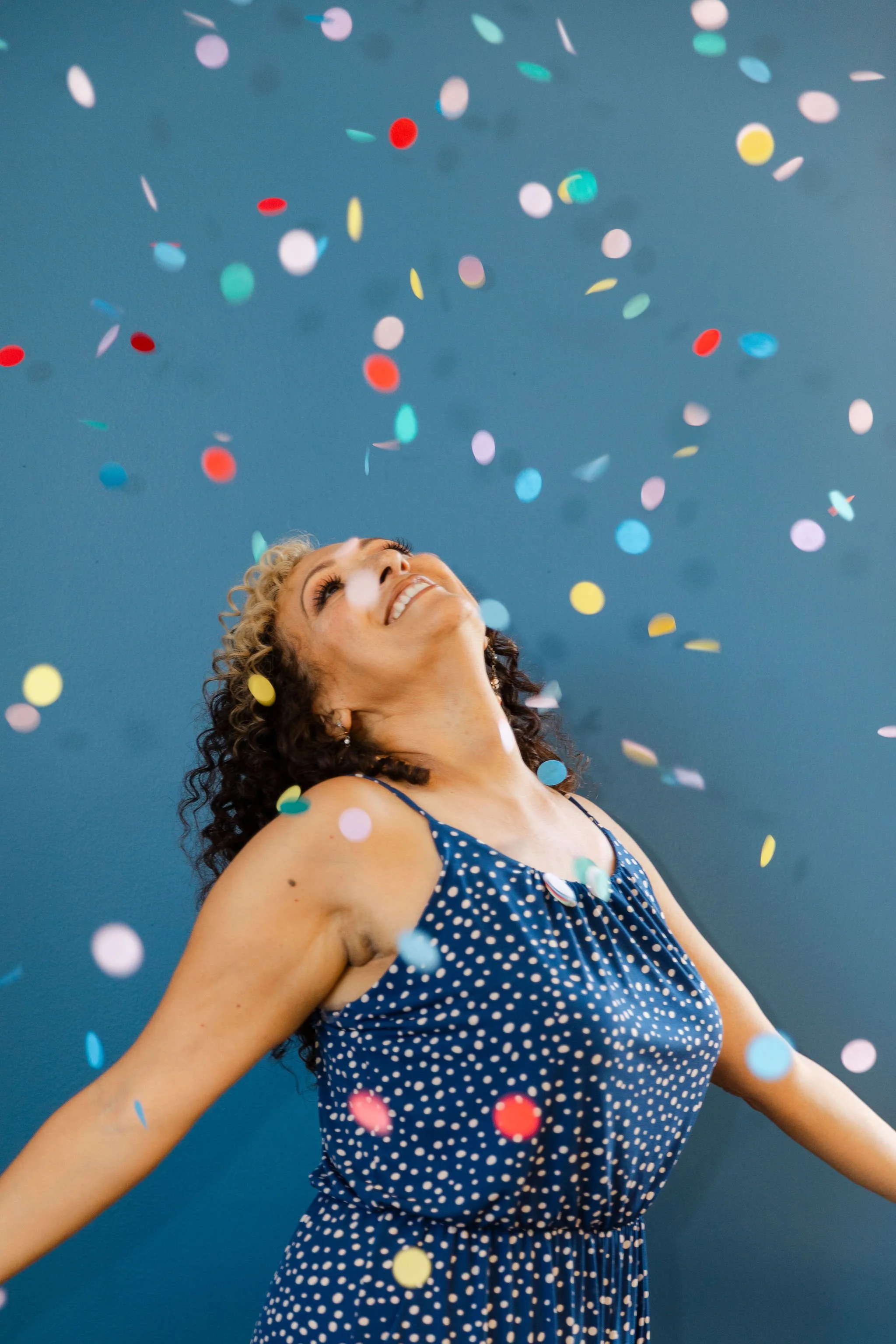 Woman in a blue polka dot dress celebrating with colorful confetti falling around her against a blue background.