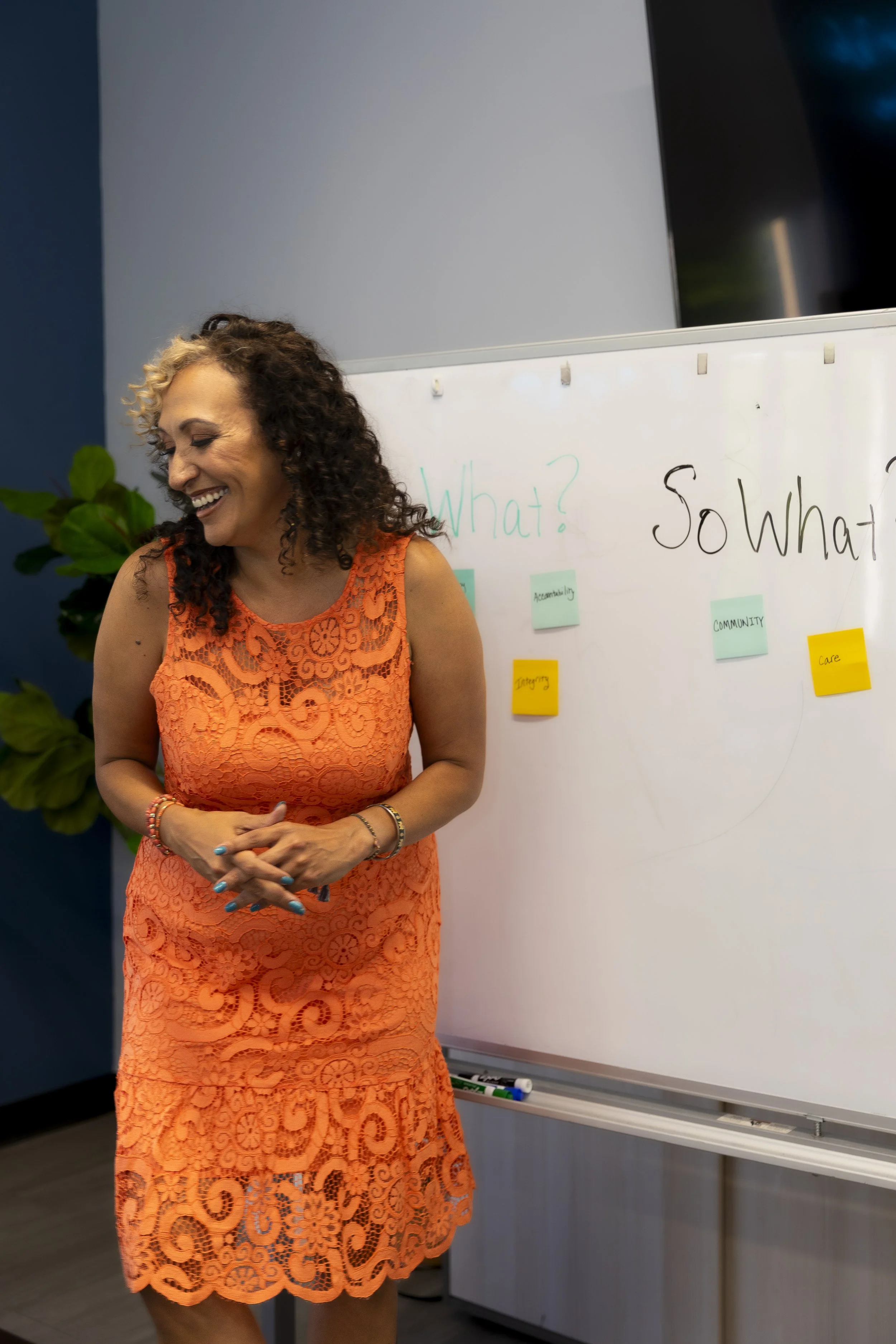 A woman with curly hair wearing an orange lace dress, standing in front of a whiteboard with written questions 'What?' and 'So what?', smiling.