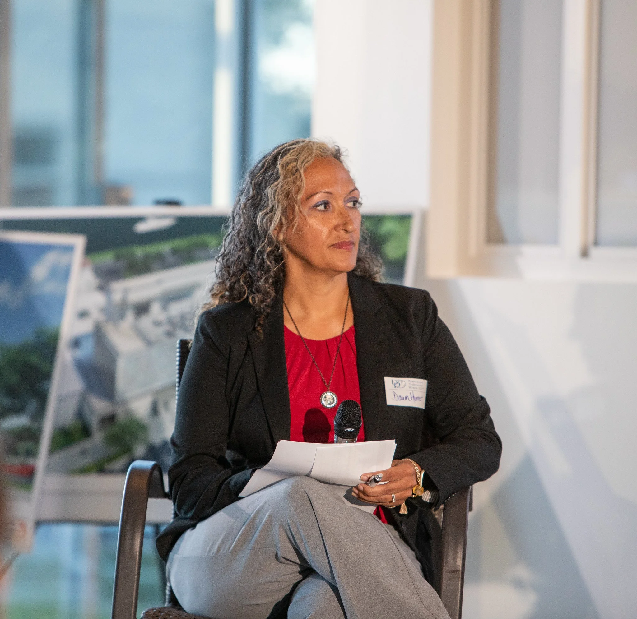 A professional woman in a black blazer and red top sitting at a table during a conference or meeting, holding a notebook and pen with a microphone in front of her, in an office setting with large windows.