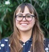 A young woman with long brown hair, glasses, and a blue polka dot shirt, smiling outdoors with green foliage in the background.