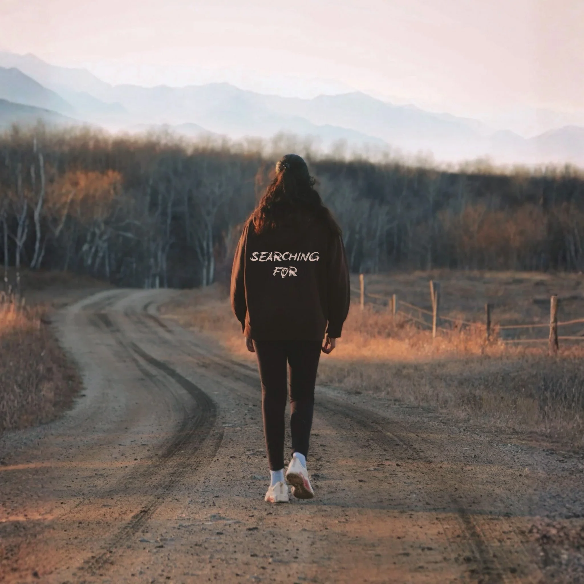 A person with long hair, wearing a black hoodie with the words "SEARCHING FOR" on it, walking down a dirt path in a rural area with bare trees and mountains in the background during sunset.