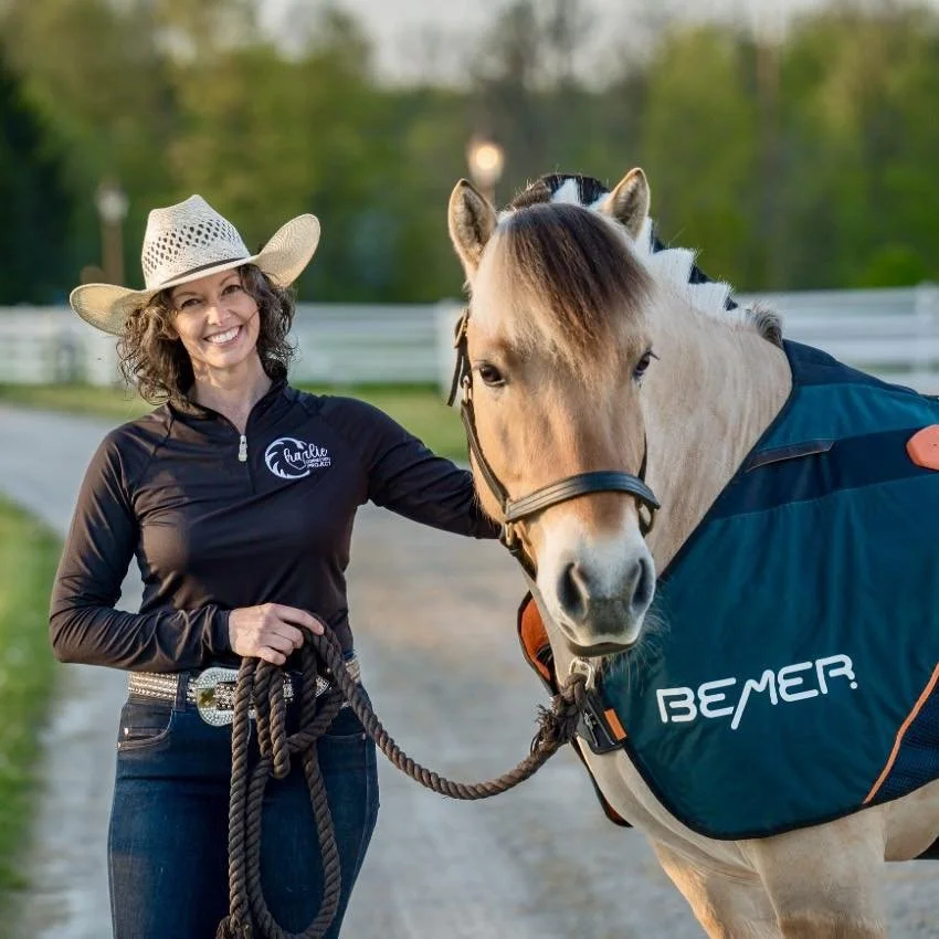 A smiling woman wearing a cowboy hat and a black jacket, holding a lead rope attached to a white horse with a blond mane, on a rural dirt path with green trees and a white fence in the background.