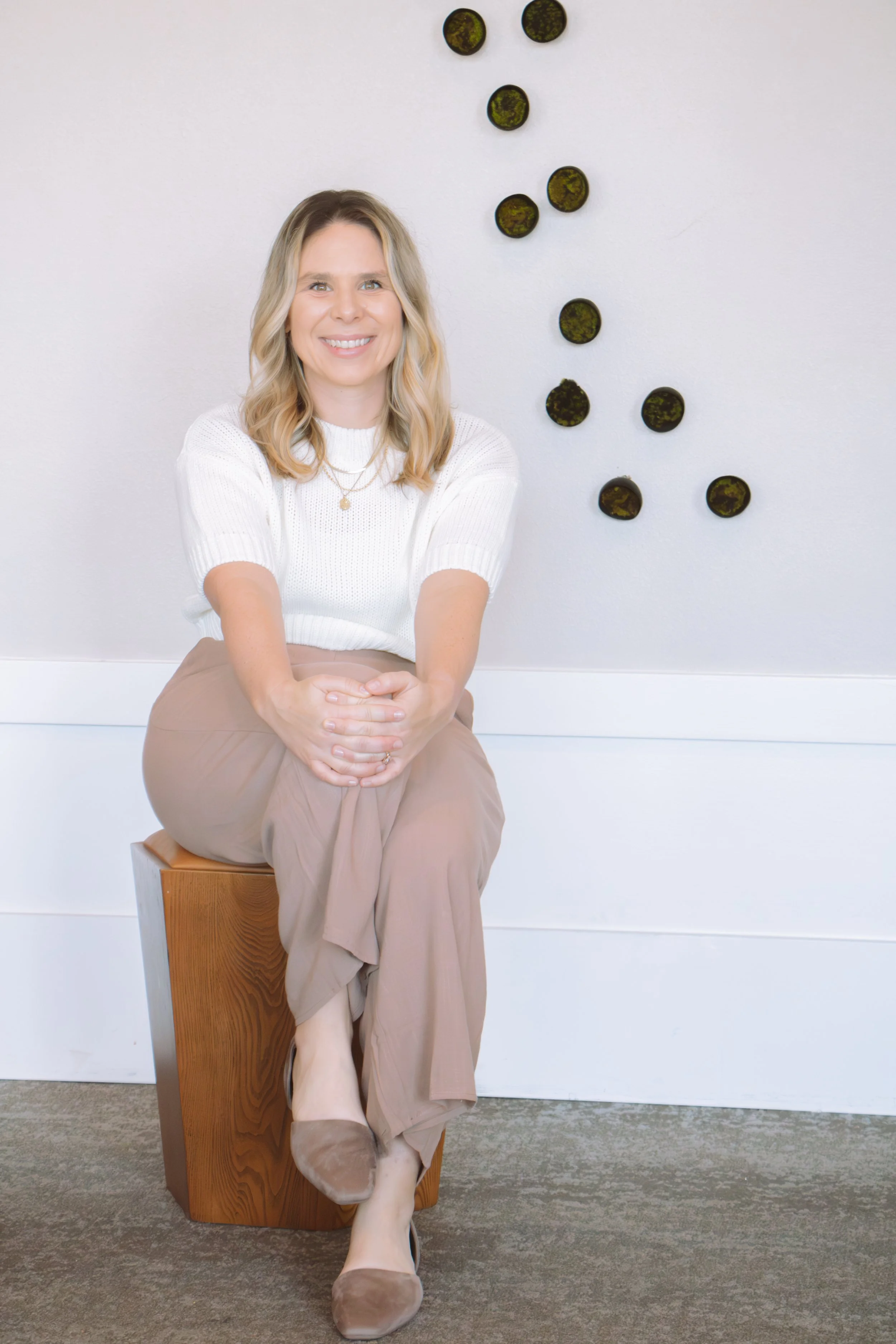 A woman with blonde hair and a white sweater sitting on a wooden block, smiling, with her hands folded in her lap. She is wearing beige wide-leg pants and beige shoes. Behind her is a white wall decorated with a scattered arrangement of small, round, greenish black objects.