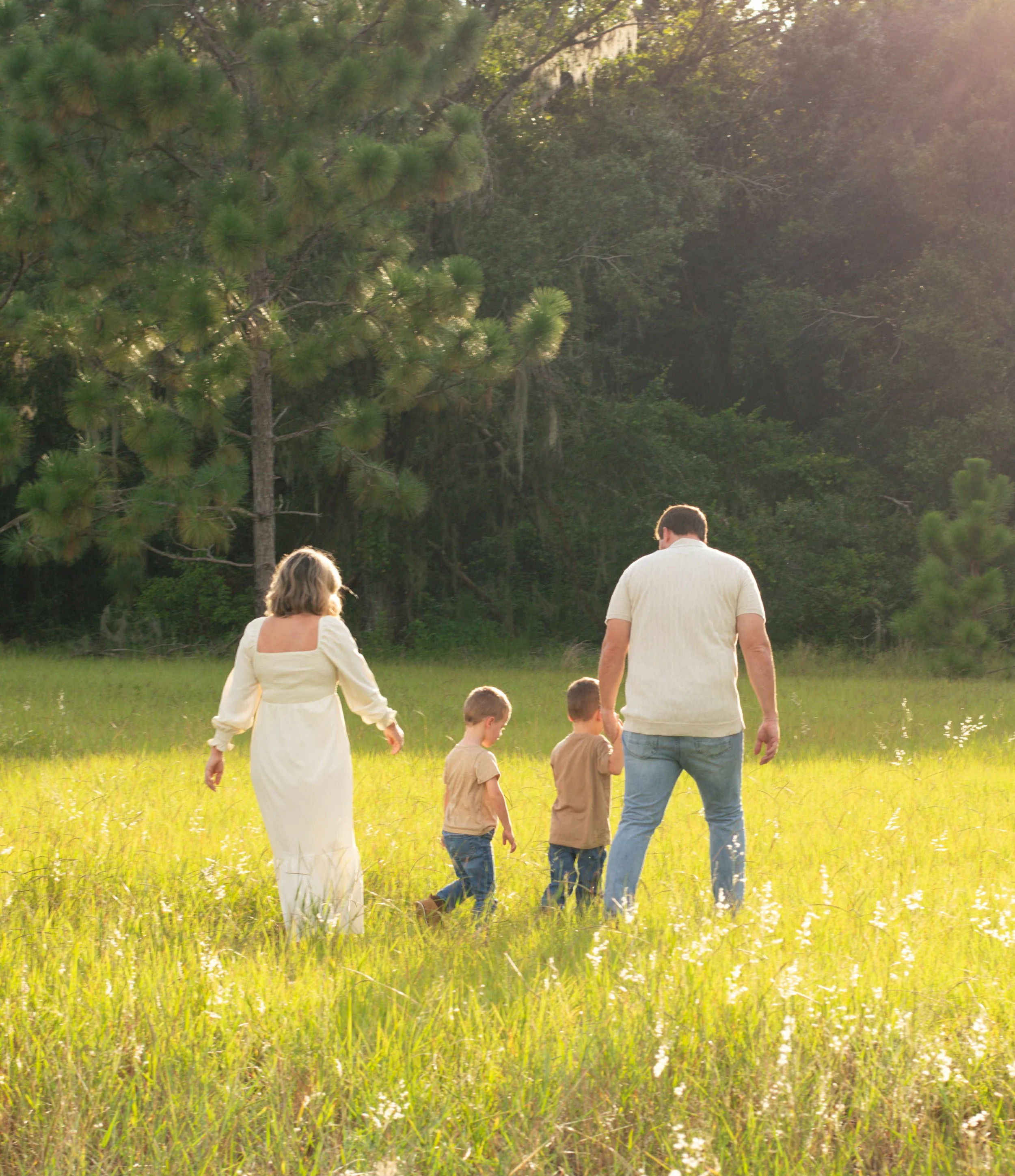A family walking through a grassy field near a wooded area with tall trees and sunlight filtering through the leaves.