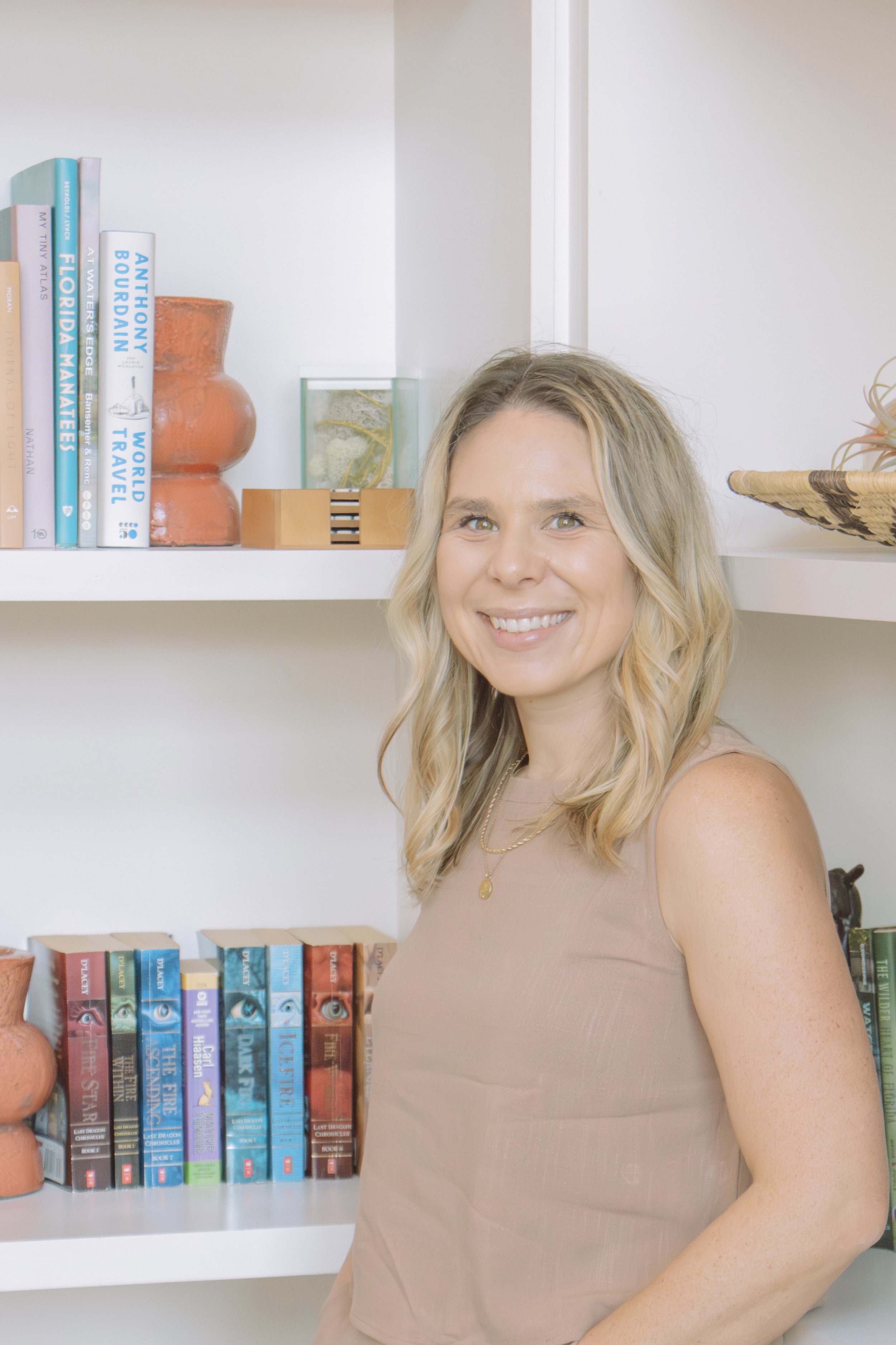 A woman with blonde hair smiling in front of a white bookshelf filled with books and decorative items, wearing a beige sleeveless top and a gold necklace.