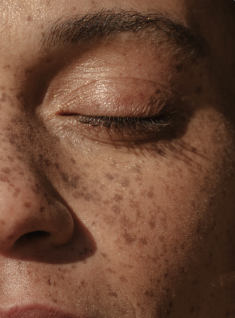 Close-up of a person's face with closed eyes, showing freckles, skin texture, and dark hair.