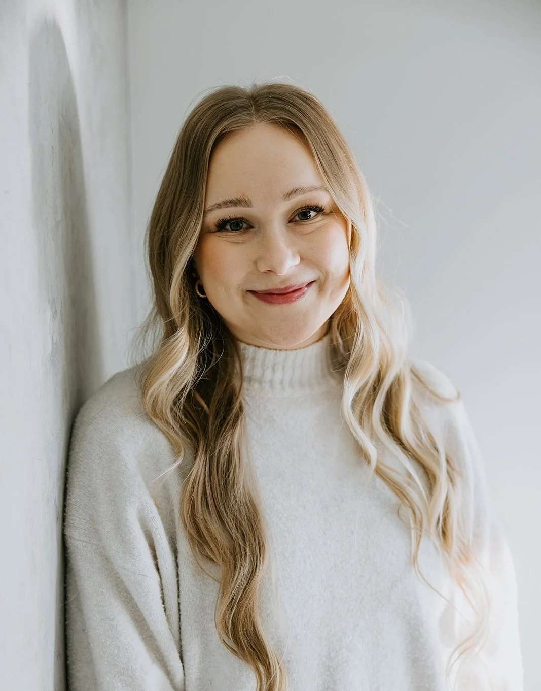 A young woman with long wavy blonde hair, wearing a cream-colored turtleneck sweater, smiling and standing against a light-colored wall.